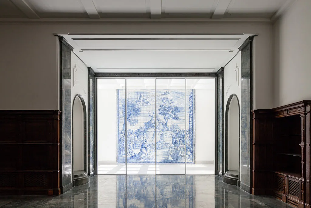 A neoclassical entryway features grey marble flooring, dark wood paneling, and arched openings looking through a glass wall to a blue and white tile mural.