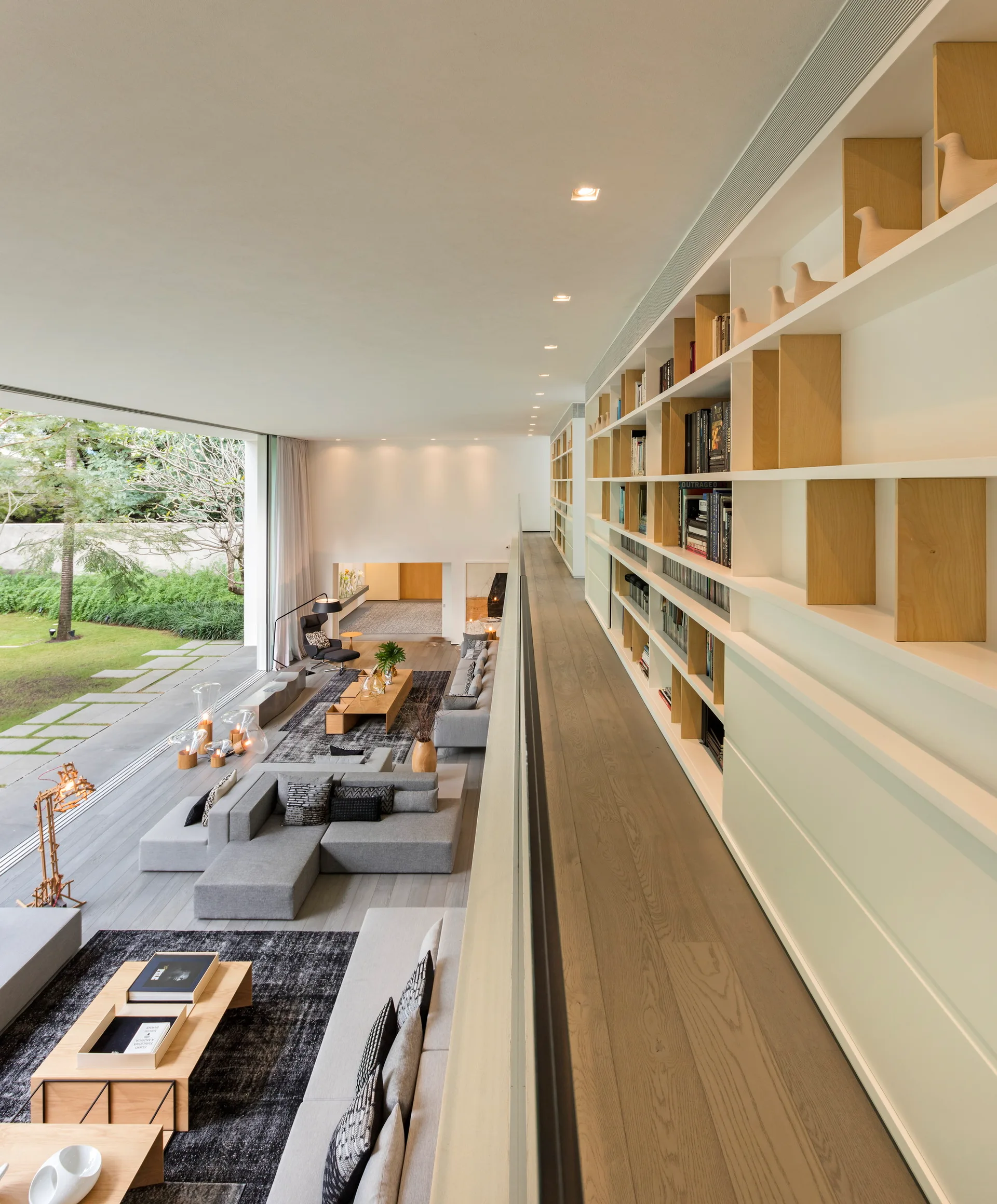 High angle view of an open-plan living room with a grey sectional sofa, wood coffee tables, and an elevated mezzanine walkway lined with white bookshelves.