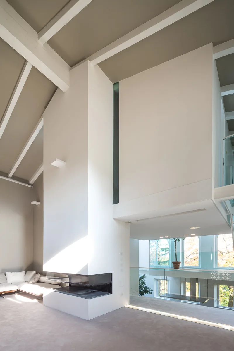 A spacious living room featuring a tall white plaster fireplace, a vaulted ceiling with white beams, and a mezzanine balcony with a glass railing.
