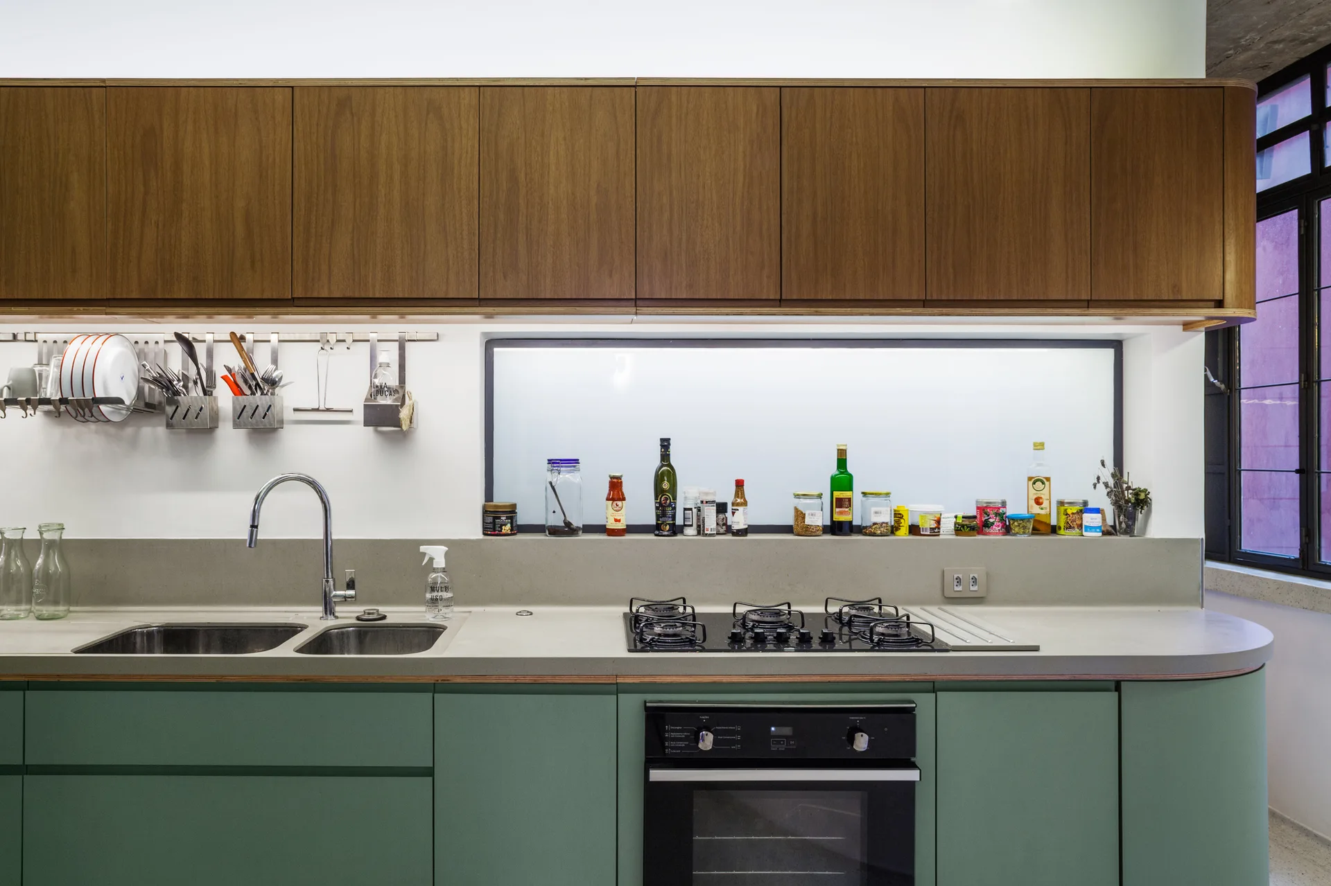 A modern kitchen with sage green lower cabinets, wood upper cabinets, a grey countertop, and a long horizontal frosted glass window.