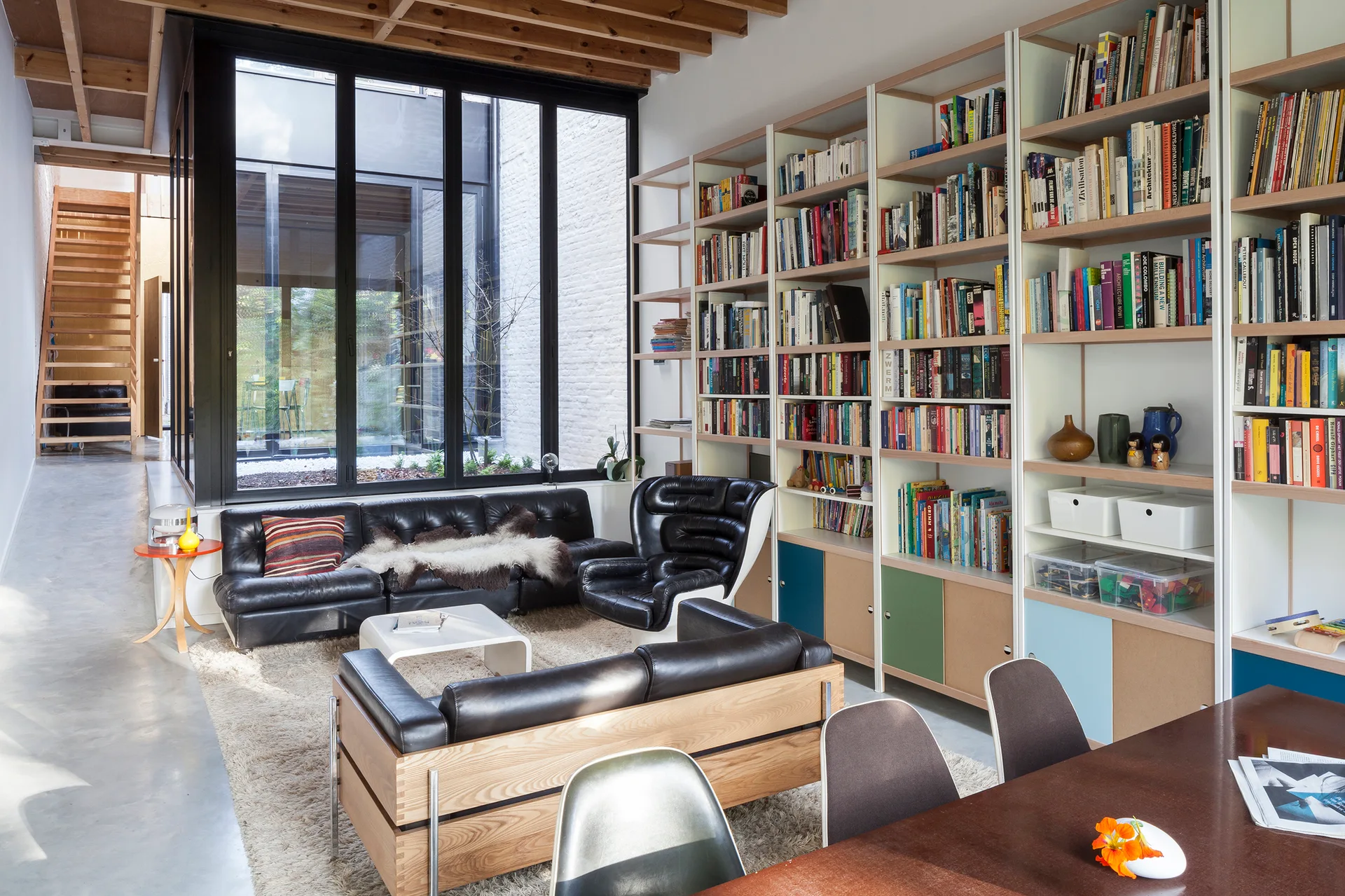 A modern living room features black leather seating on a shag rug, a massive floor-to-ceiling bookshelf, and exposed wooden ceiling beams.