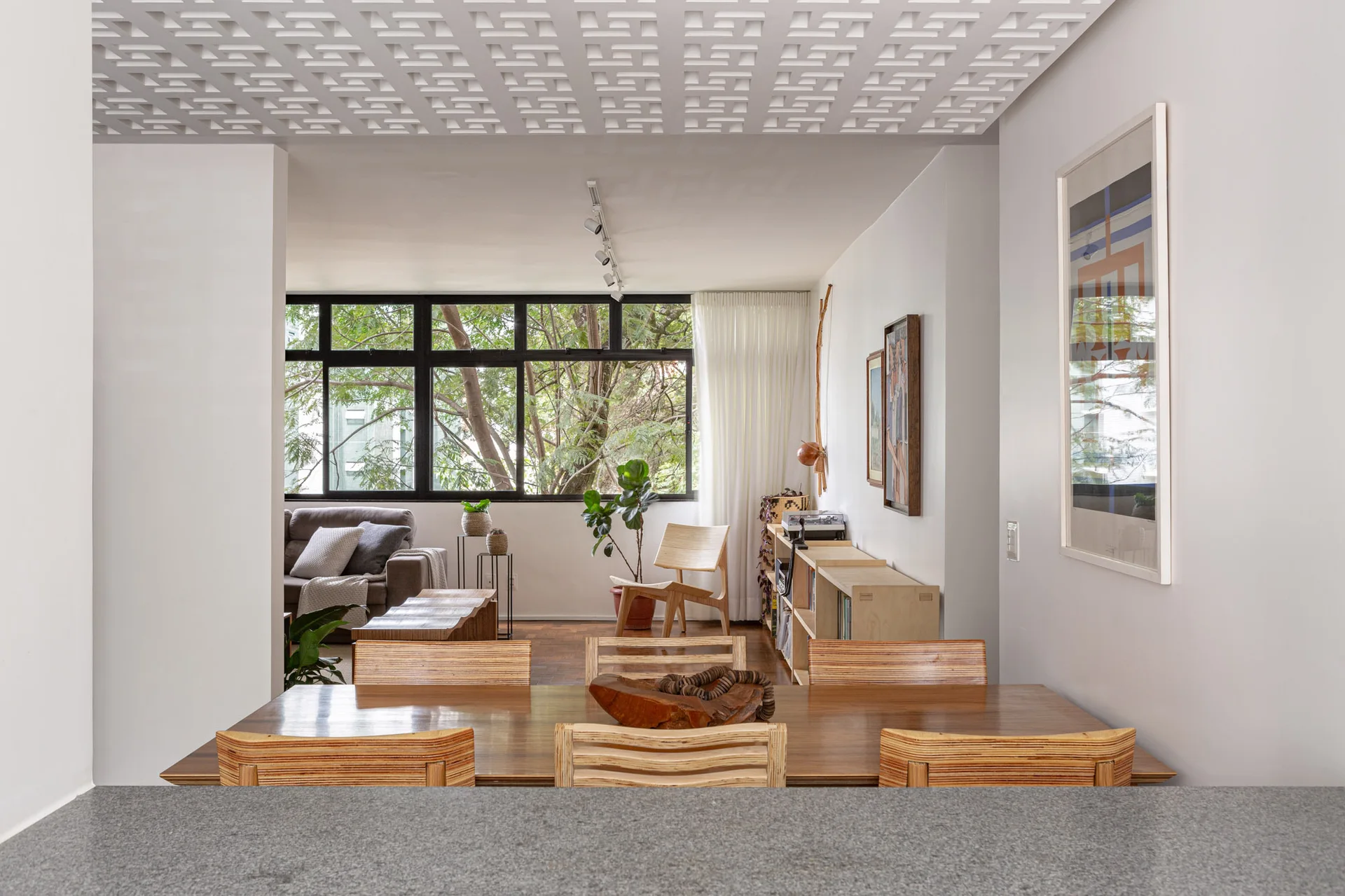 An open dining space with a wooden table and chairs features a geometric white grid ceiling and a large window overlooking trees.