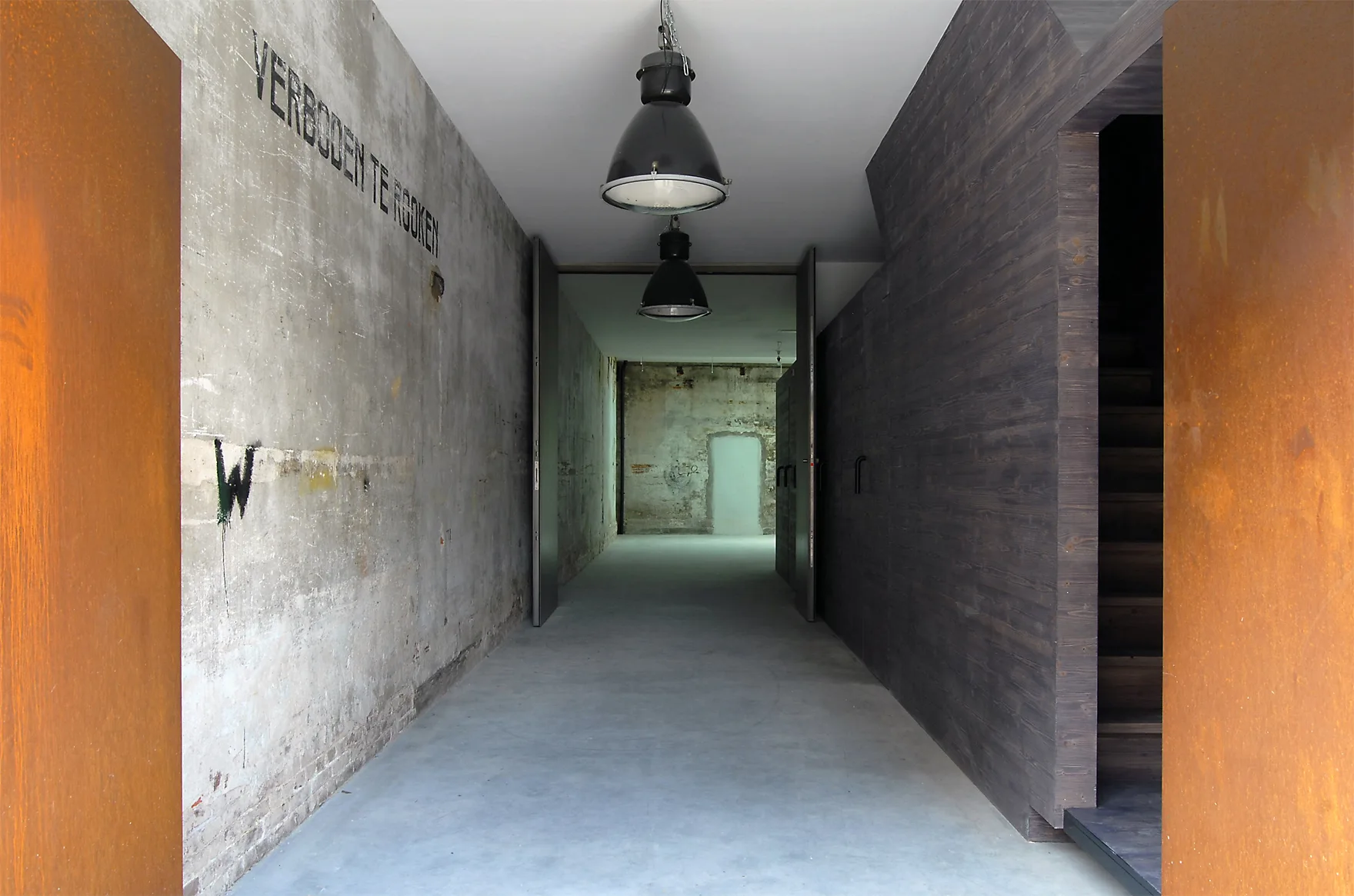 A long industrial hallway features raw concrete walls, rusted steel panels in the foreground, dark wood paneling, and black pendant lights.