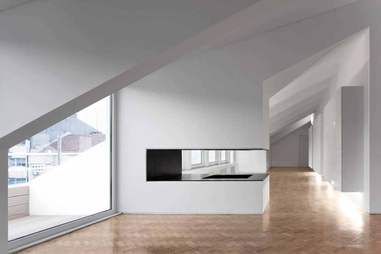 A minimalist white attic room featuring sloped ceilings, light wood herringbone flooring, a large window, and a built-in black partition fireplace.