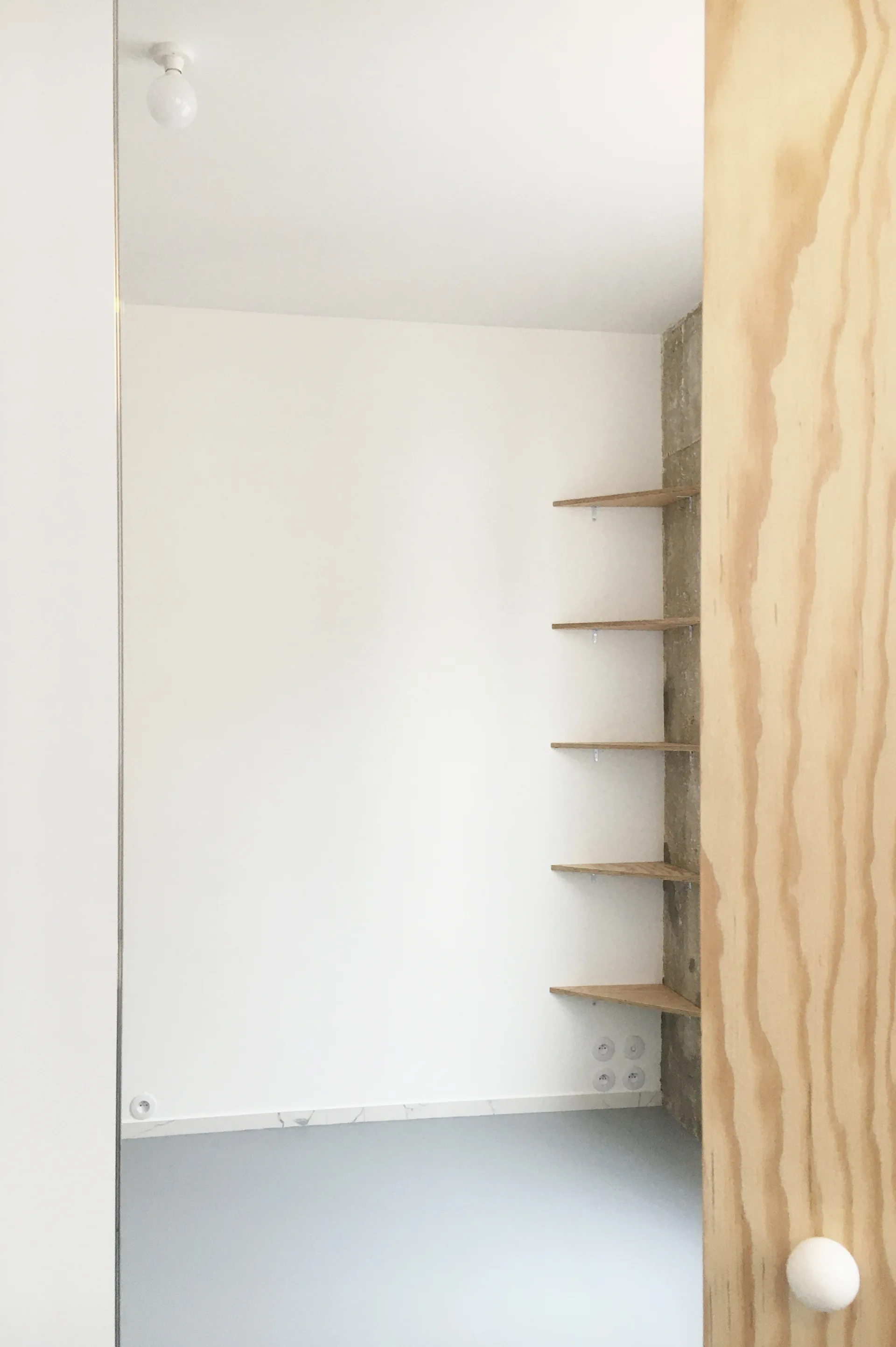A minimalist room corner features floating triangular plywood shelves mounted on a vertical strip of raw concrete next to plain white walls.