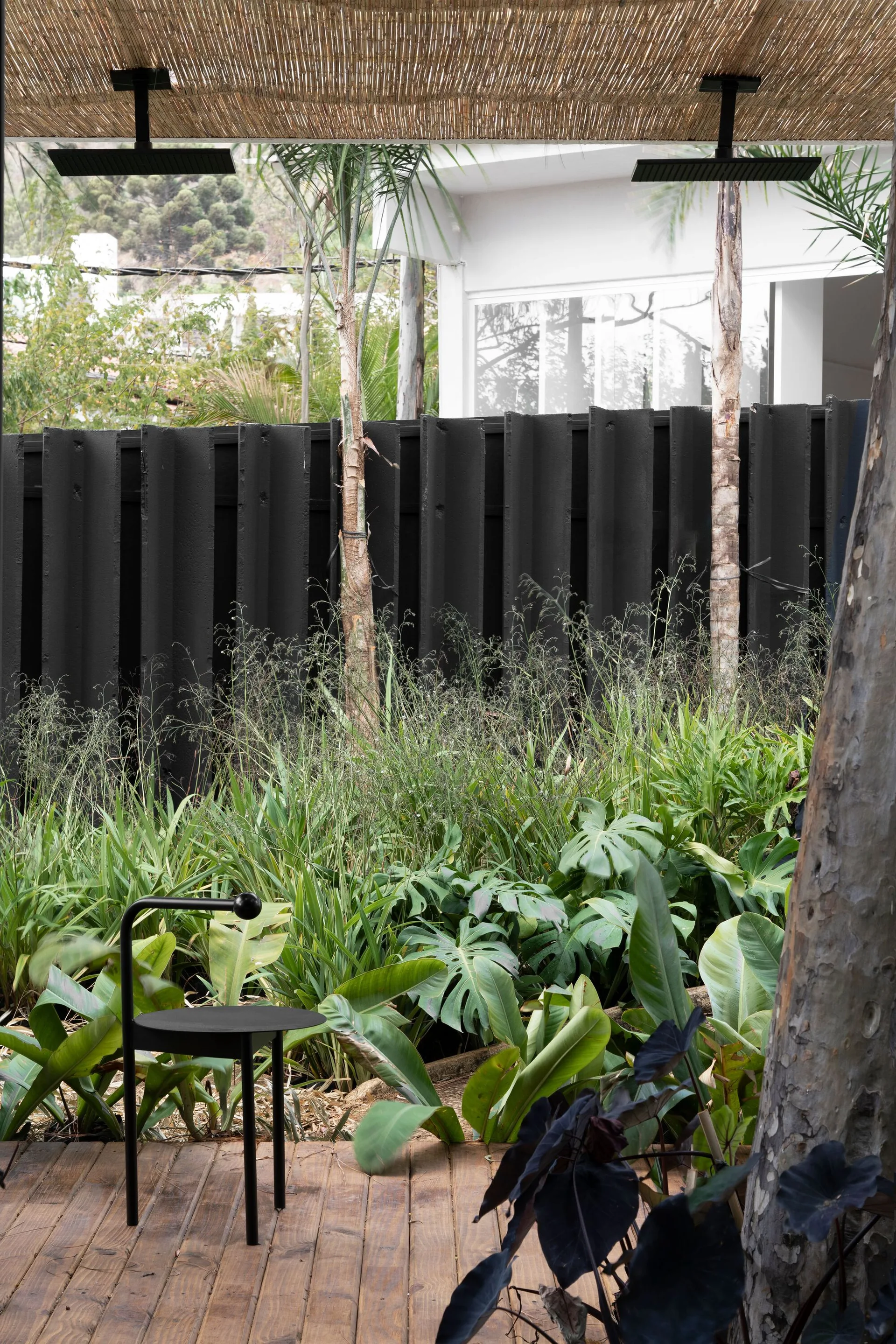 A black minimalist metal stool sits on a wooden deck next to lush tropical plants and a black textured slat fence under a bamboo awning.