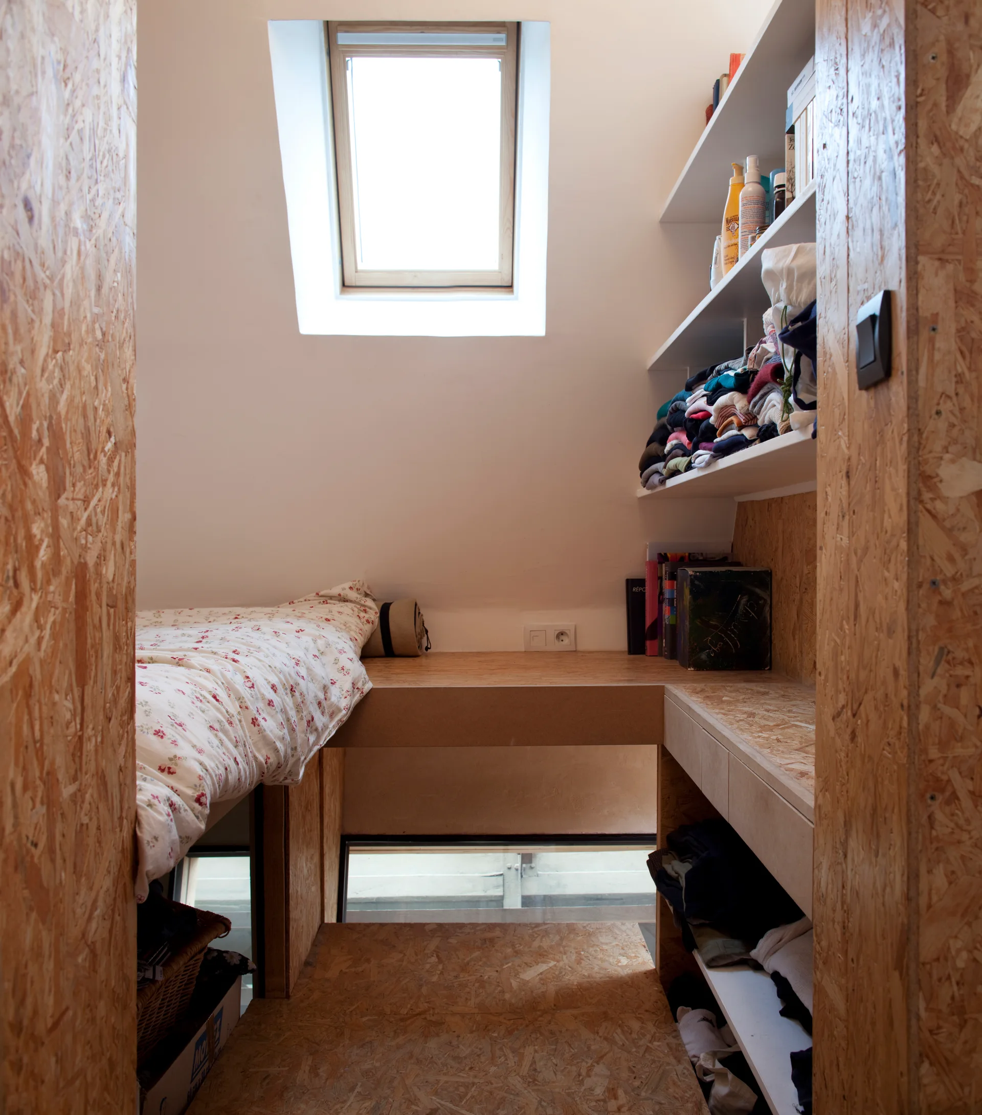 A small attic bedroom features a custom oriented strand board platform bed, open wall shelves, and a single skylight.