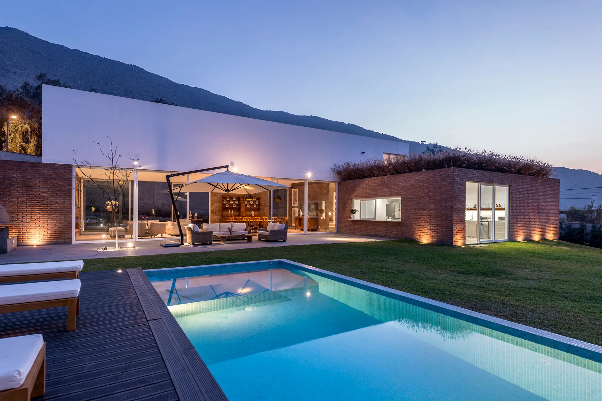 A modern residential exterior at twilight features a rectangular swimming pool, wooden deck, textured brick facade, and large glass windows.