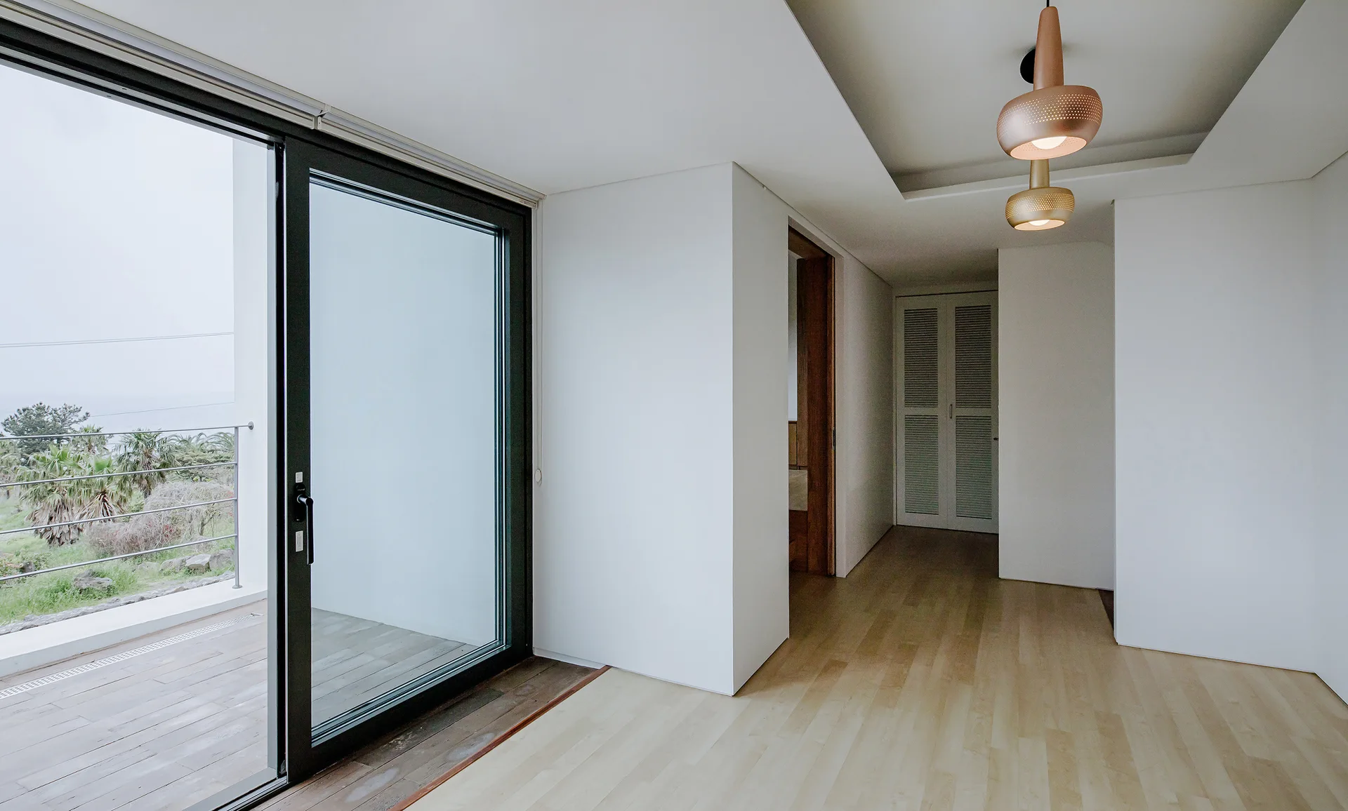 A bright minimalist hallway with light wood floors features a large sliding glass door opening to a deck and a copper pendant light.
