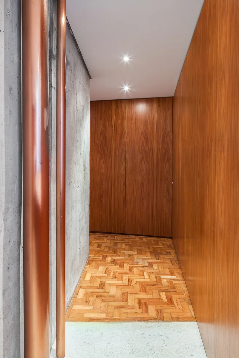 A modern corridor shows vertical copper pipes next to a raw concrete wall opposite warm wood paneling and a herringbone parquet floor.