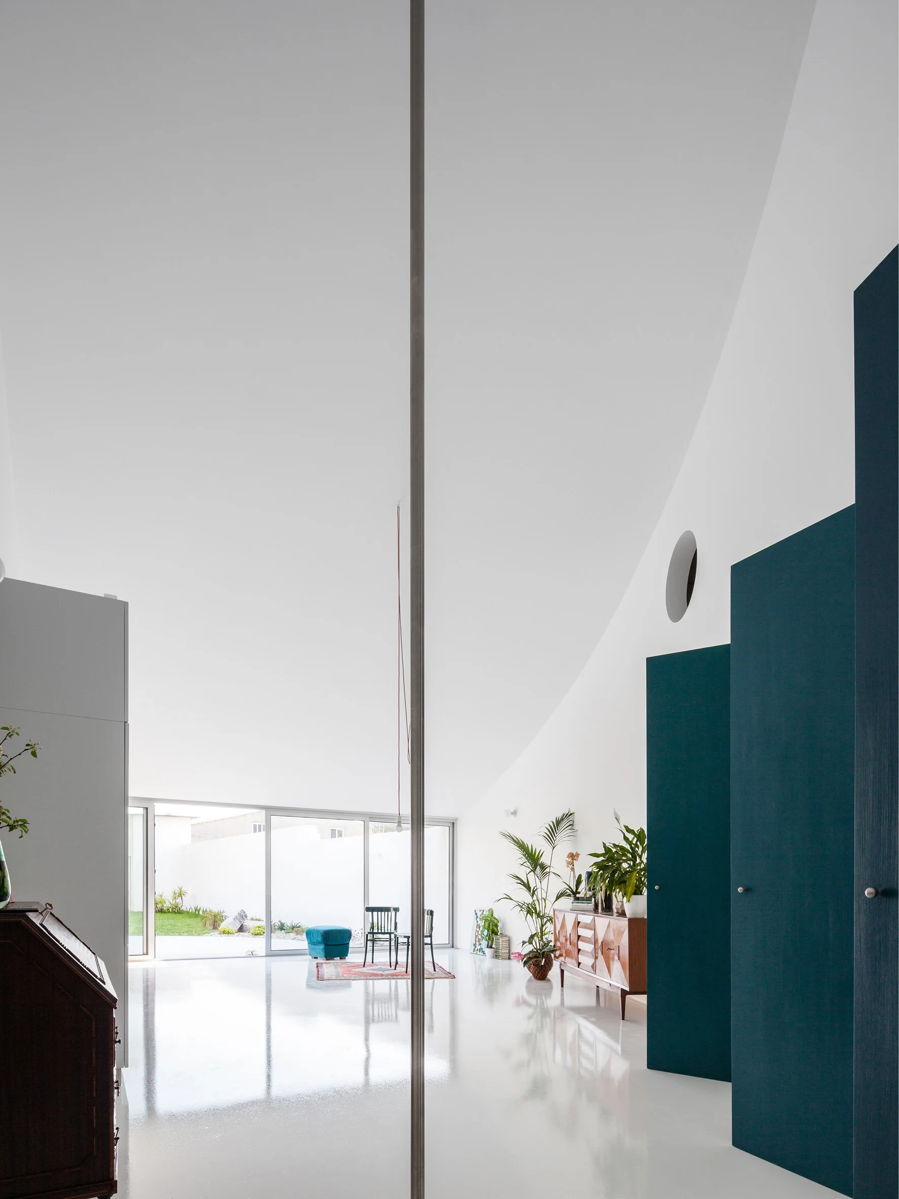 A minimalist interior features a dramatically slanted ceiling, high-gloss white floors, teal cabinets, and a glass wall leading outside.