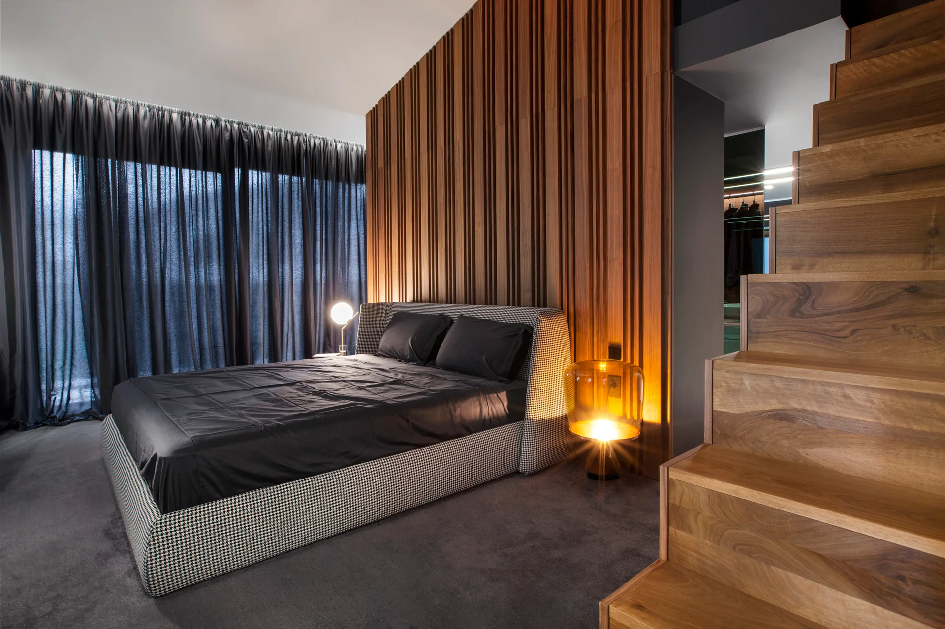 A modern bedroom with a houndstooth patterned bed set against a vertical wood slat wall next to a wooden staircase.