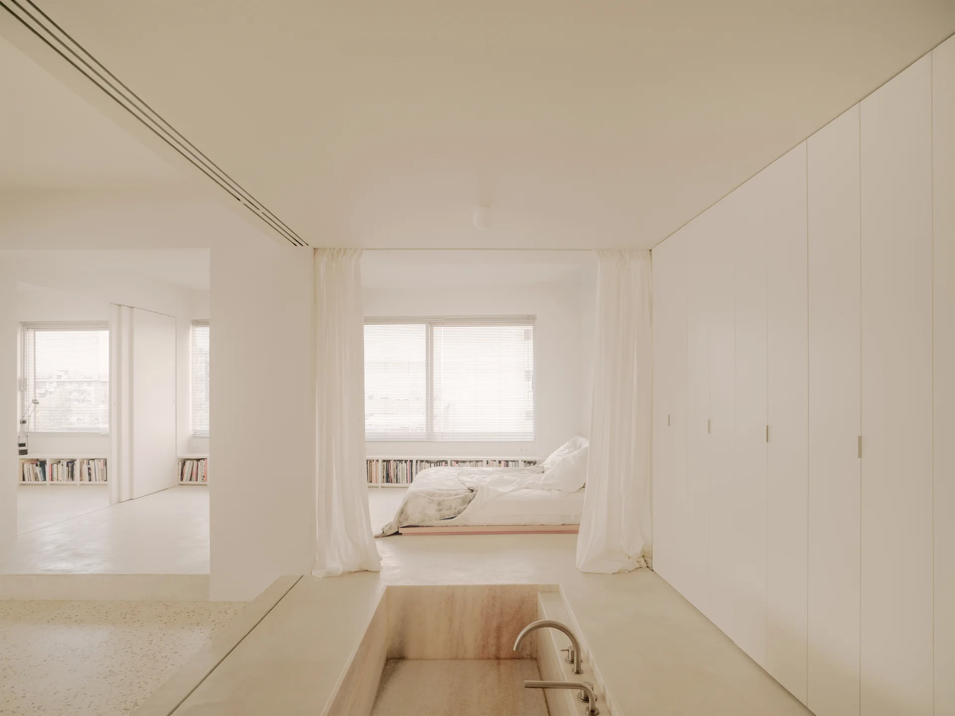 A minimalist open-plan suite features a stone sunken bathtub in the foreground and a bedroom divided by sheer white curtains in the background.