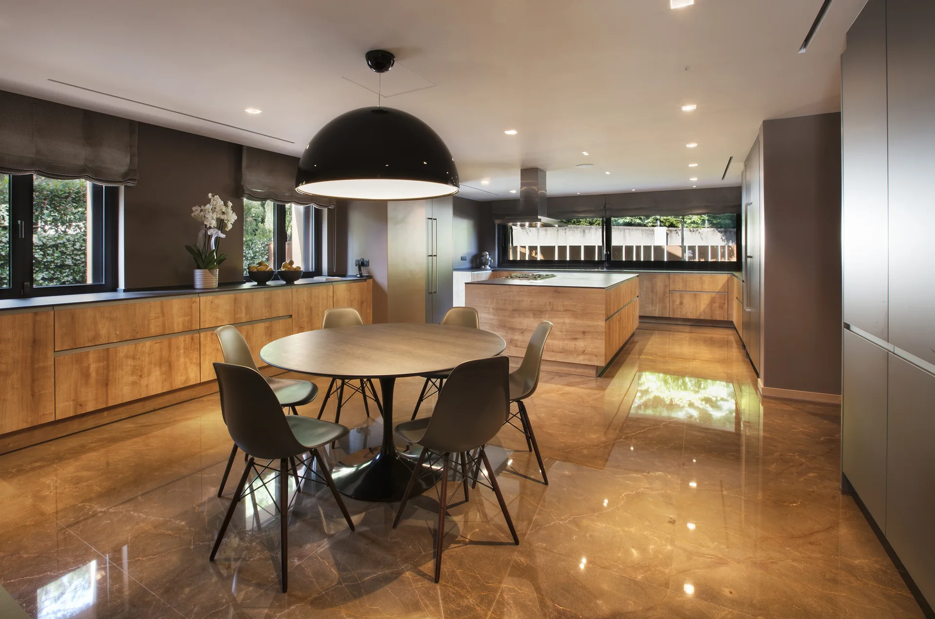 A modern open kitchen and dining area with warm wood cabinets, highly reflective brown floor tiles, and a round black table.