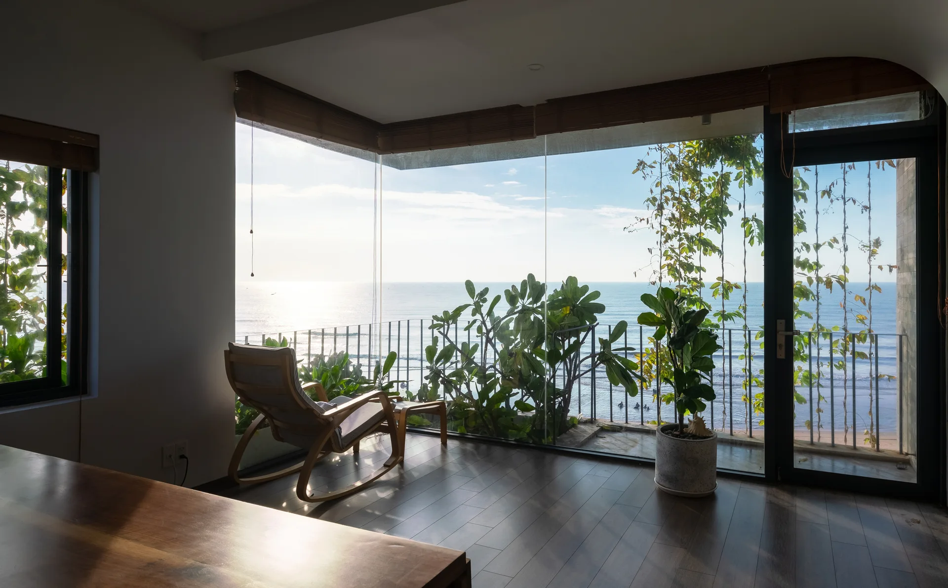A wooden rocking chair sits on dark wood floors facing expansive floor-to-ceiling corner windows with a panoramic ocean view and balcony.