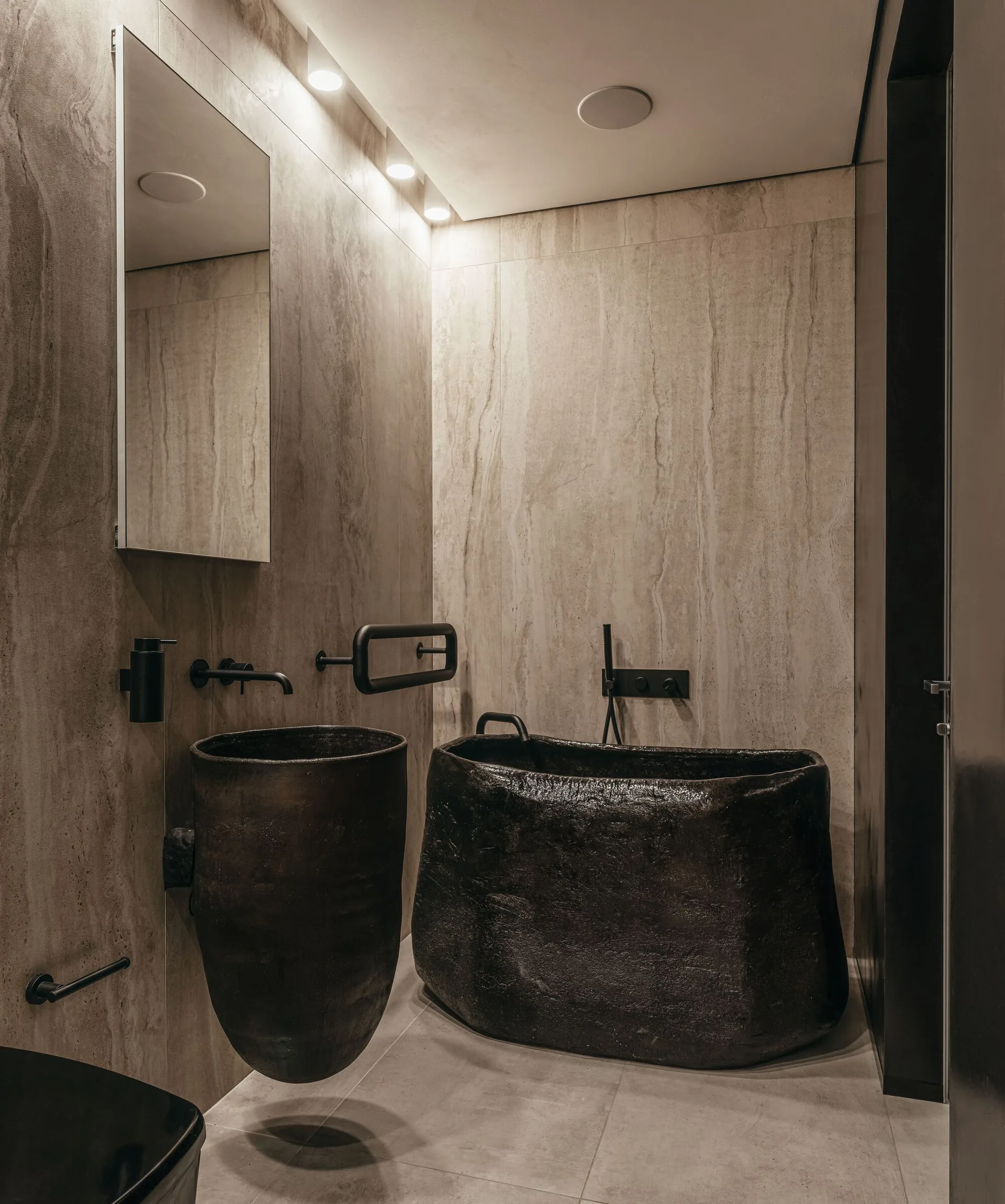 A modern bathroom featuring a dark, textured freestanding bathtub, a matching pedestal sink, and veined stone wall panels under recessed spotlights.
