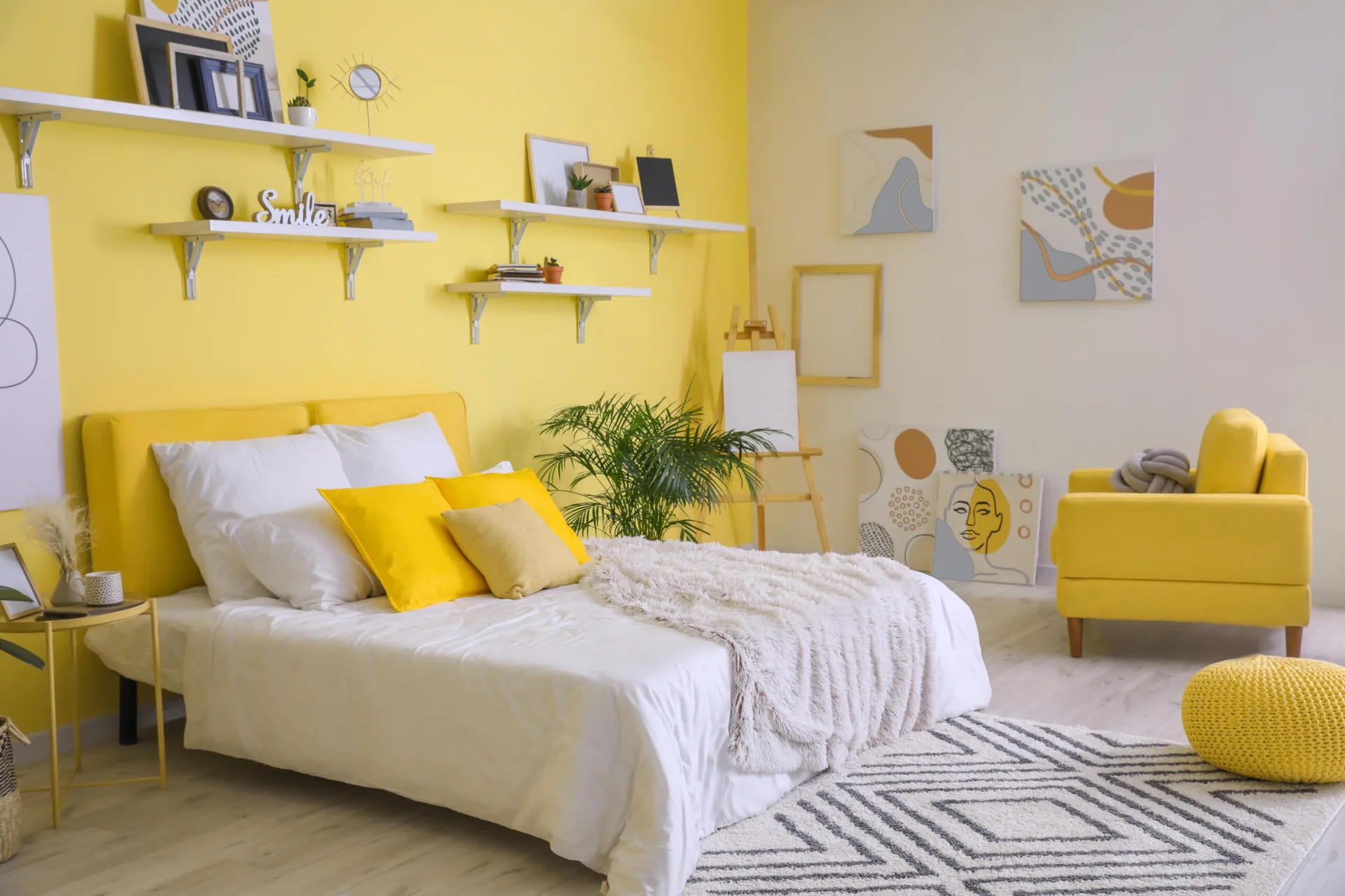 A bright bedroom featuring a yellow accent wall, a matching yellow upholstered bed, white floating shelves, and a yellow armchair on light flooring.