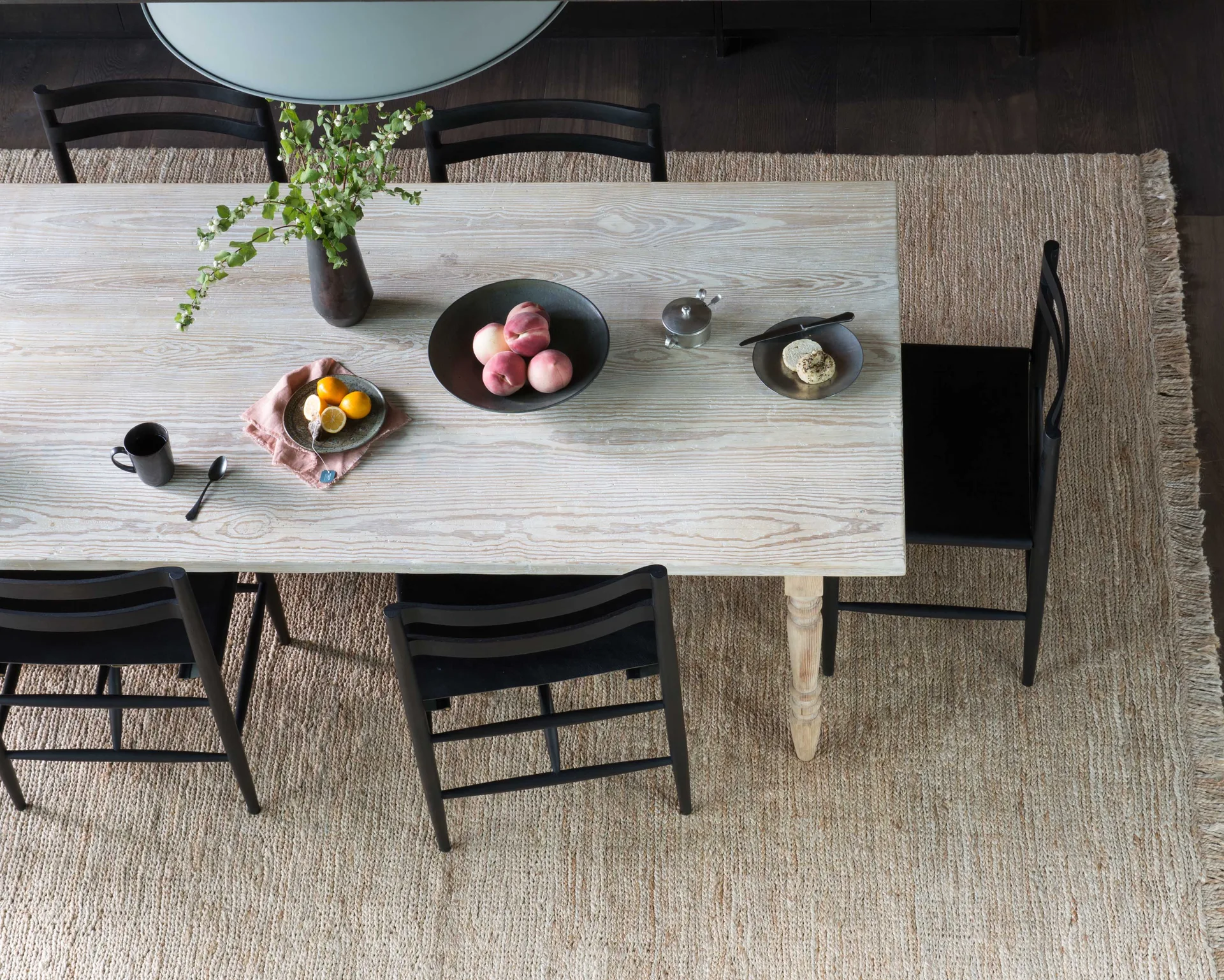 An overhead view of a light-washed wood dining table paired with black spindle-back chairs on a textured woven jute rug.