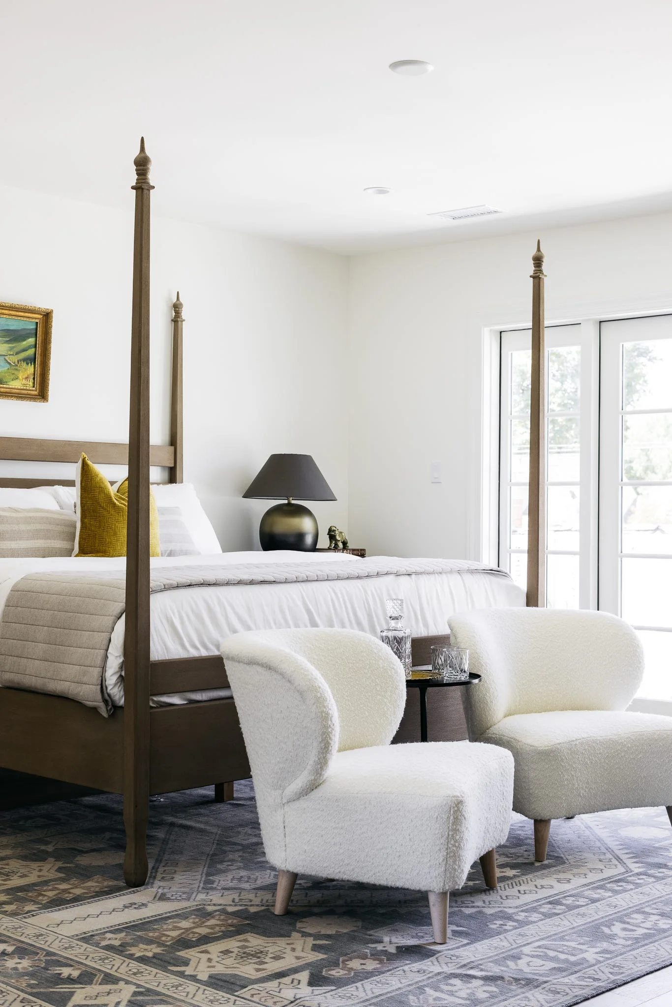 A bright bedroom featuring a wooden four-poster bed, two white boucle accent chairs with a small side table, and a patterned rug.