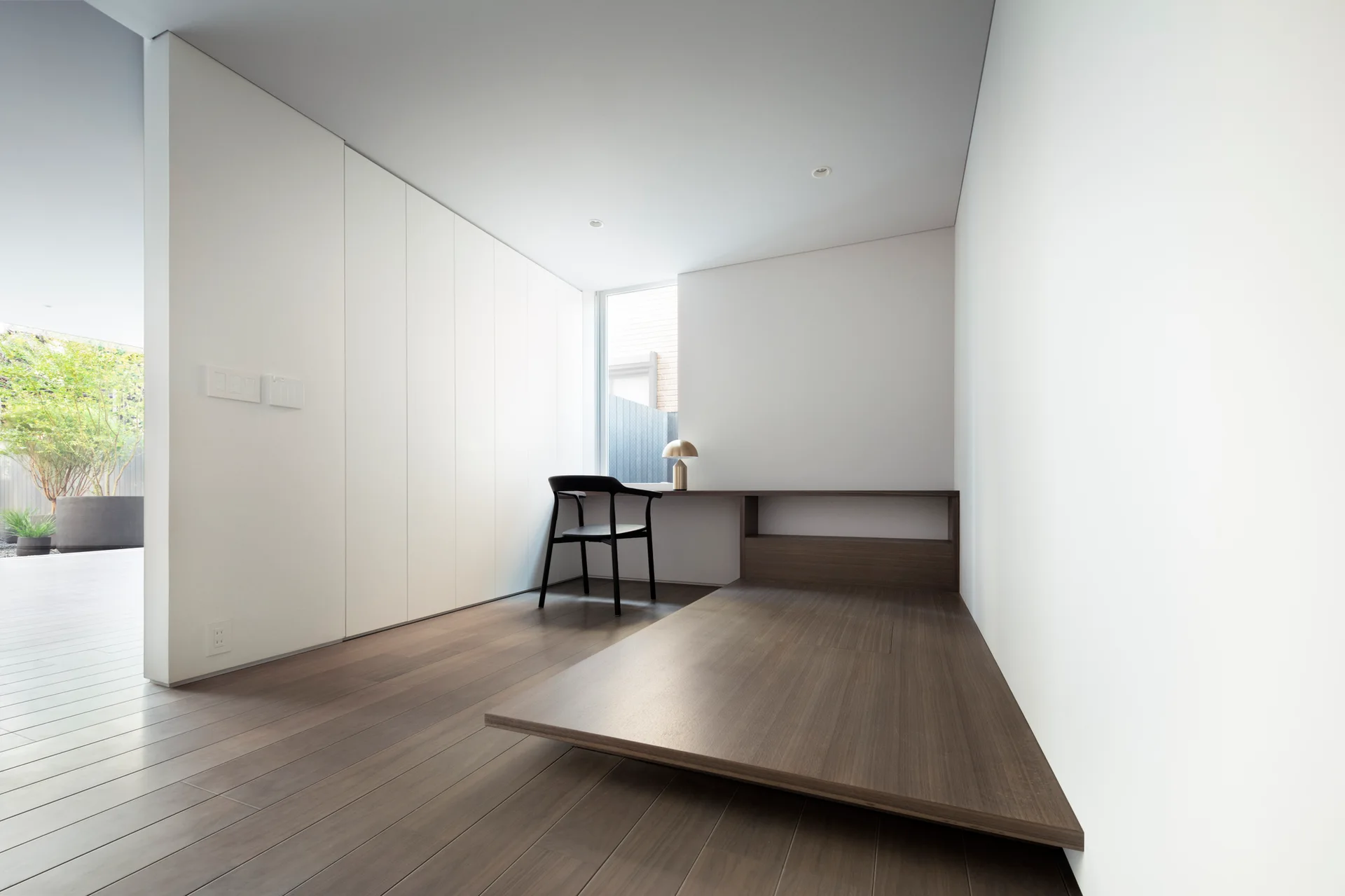 A minimalist workspace featuring a raised dark wood platform integrated with a desk, a black wood chair, a gold lamp, and tall white storage cabinets.