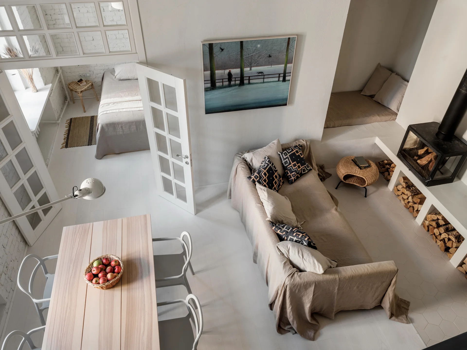 High-angle view of a white living area featuring a linen-covered sofa, a dining table, and a built-in fireplace with stacked wood underneath.