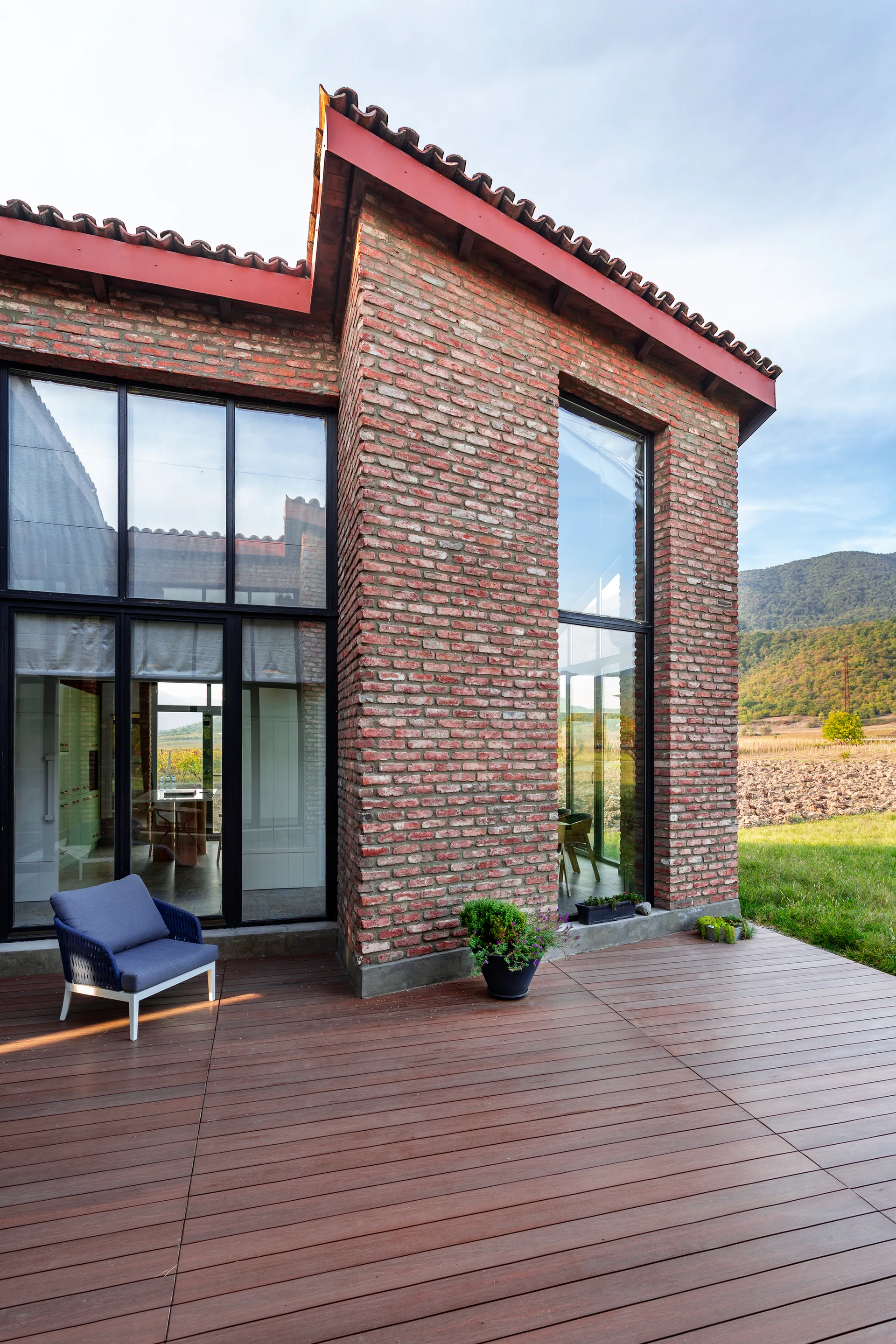 A modern rustic red brick home features large glass windows and a wide wood plank deck with a blue lounge chair.