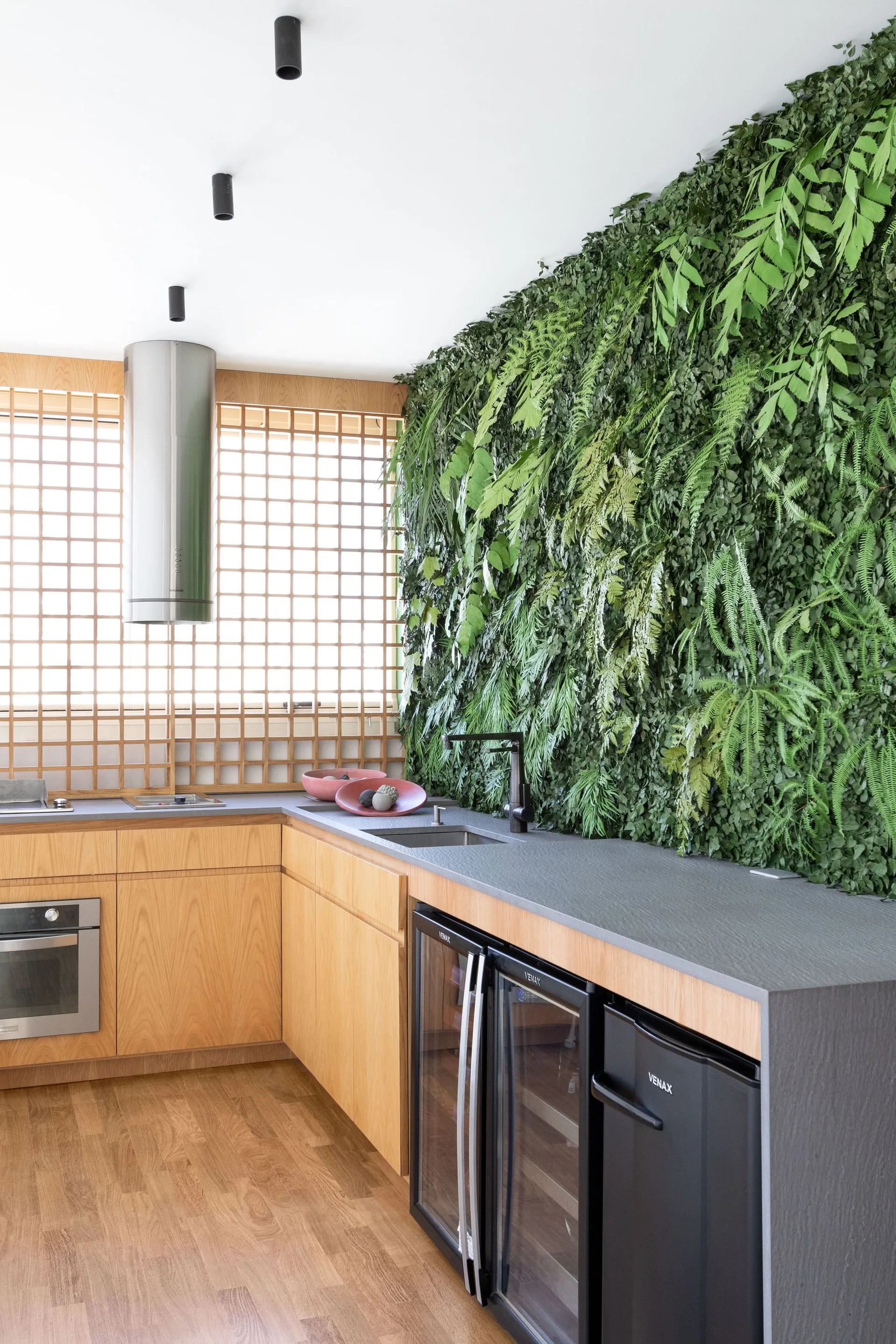 A modern kitchen featuring warm wood cabinetry, a dark grey countertop, an integrated sink, a grid wooden window screen, and a lush vertical green wall.