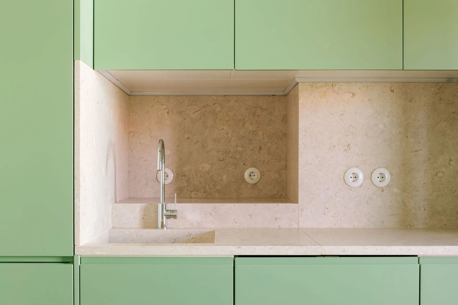 A close-up of sage green cabinets featuring an integrated beige stone sink basin, matching stone backsplash, and a modern chrome faucet.