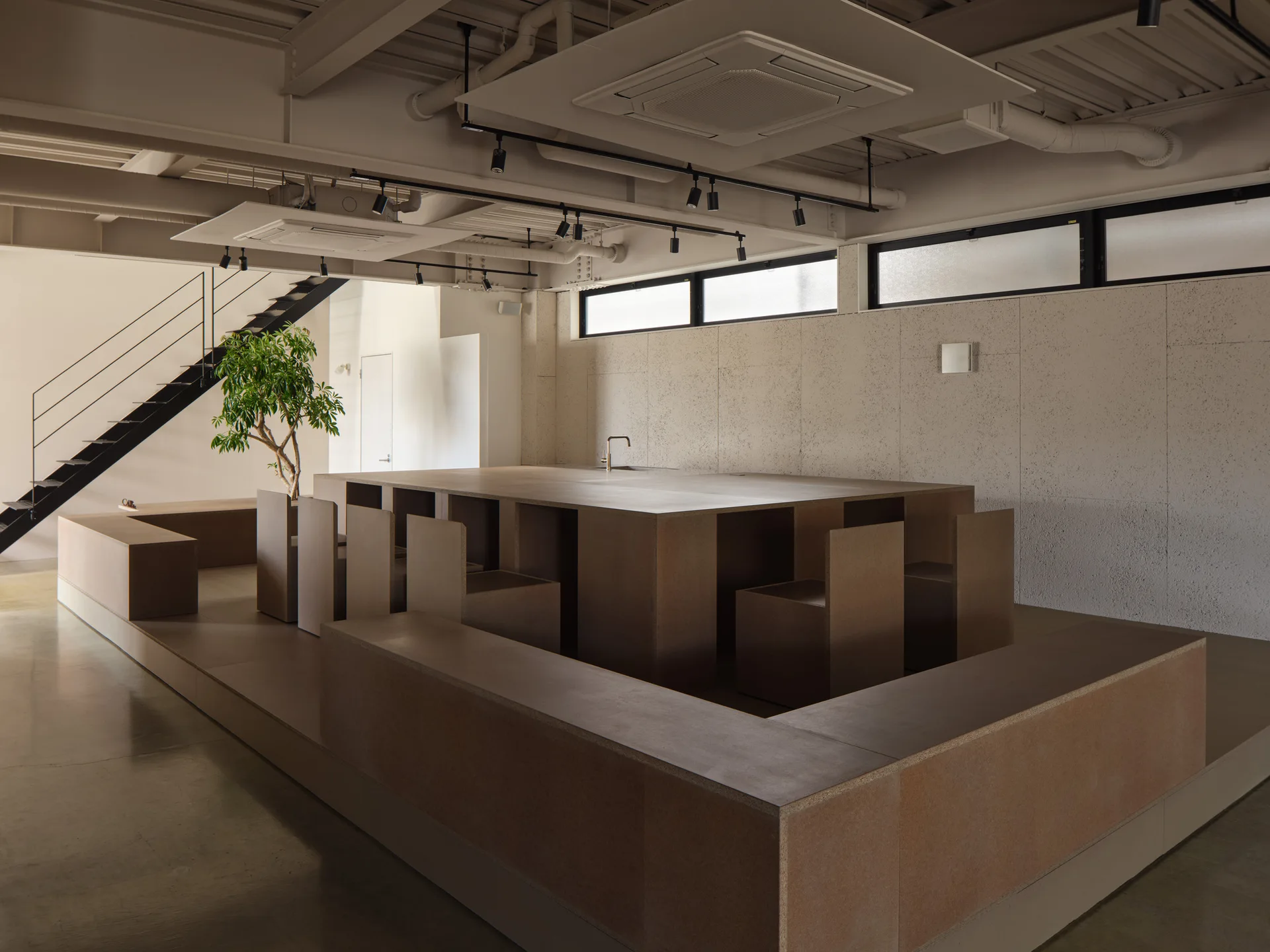 A large tan monolithic kitchen island with built-in bench seating and block chairs is set in an industrial loft space with an exposed ceiling.