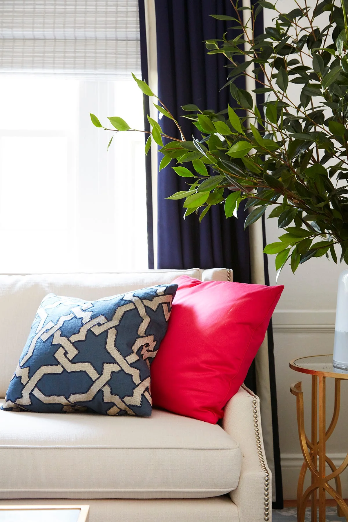 A cream sofa features a geometric navy pillow and a solid hot pink pillow next to a gold side table holding a leafy green arrangement.