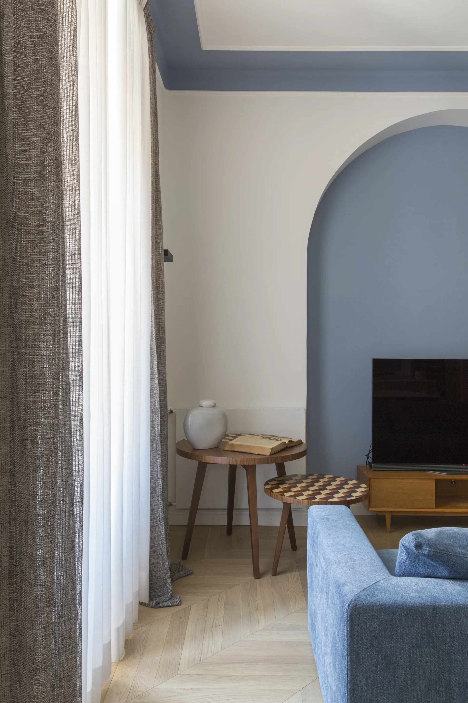A modern living room featuring a blue painted arched alcove, a blue fabric sofa, nested wooden side tables, and herringbone wood flooring.