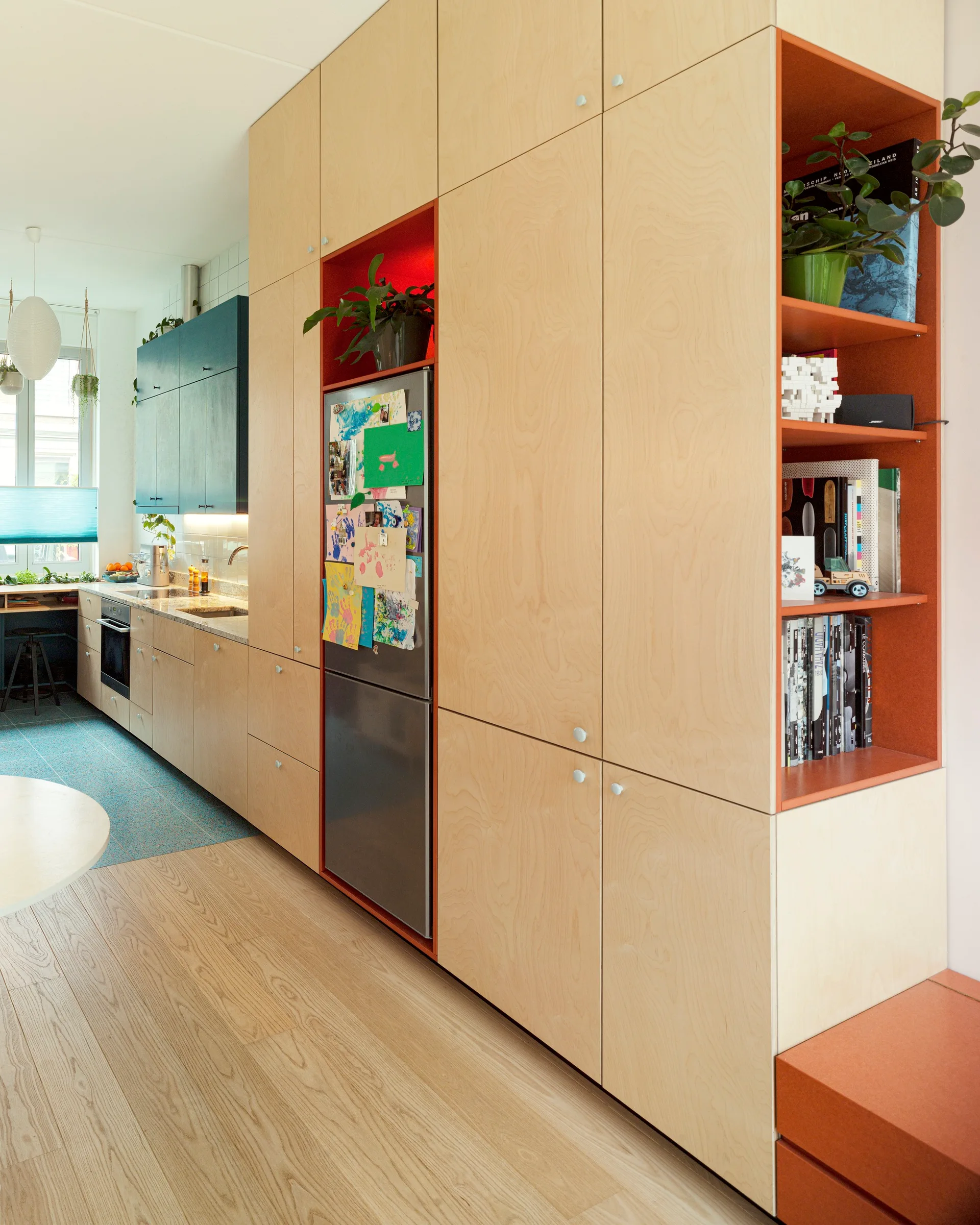 A modern Scandinavian kitchen features light-colored plywood cabinets with built-in orange shelves and an integrated stainless steel refrigerator.