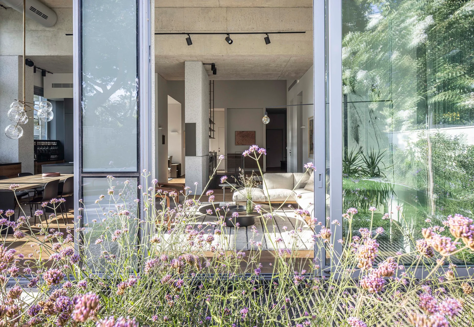 A modern living room with a white sectional sofa and exposed concrete ceiling is viewed through large sliding glass doors framed by wildflowers.