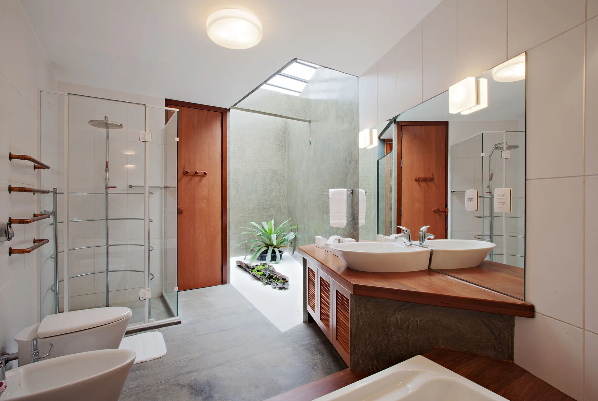 A modern bathroom with a wooden vanity, a vessel sink, a glass-walled shower, and an indoor courtyard under a skylight.