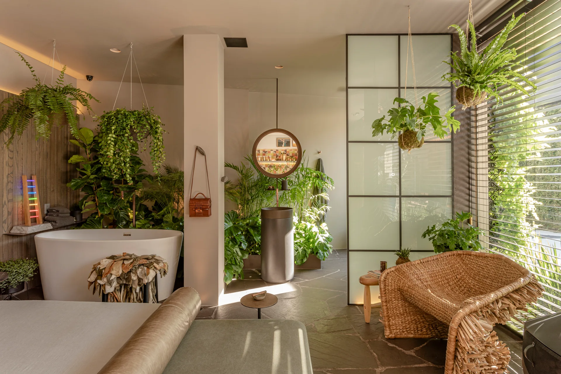 A nature-inspired bathroom features a freestanding white bathtub, a pedestal sink, a frosted glass room divider, and lush hanging plants.