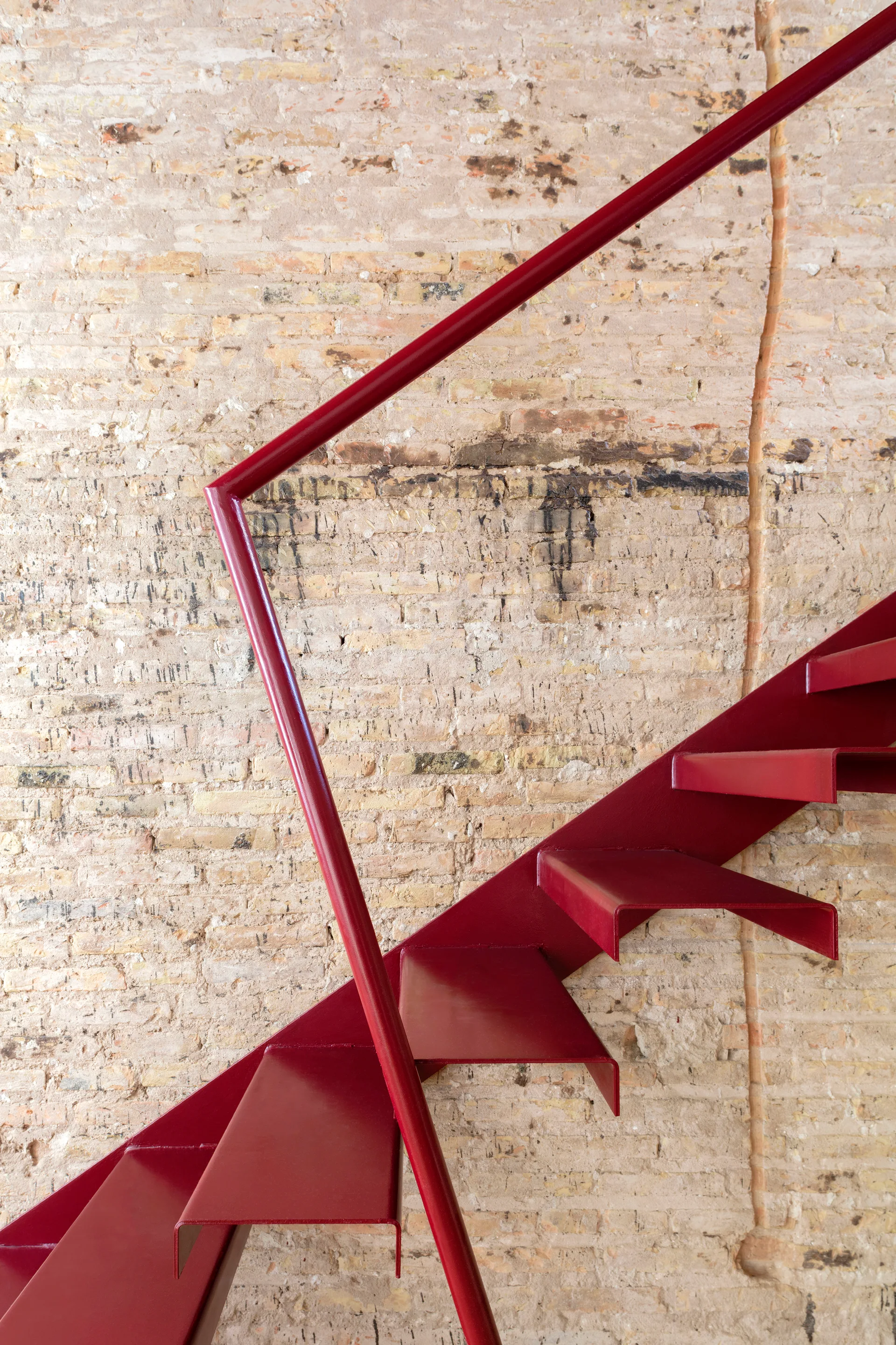 A red metal staircase with a matching railing is mounted against a textured beige exposed brick wall.