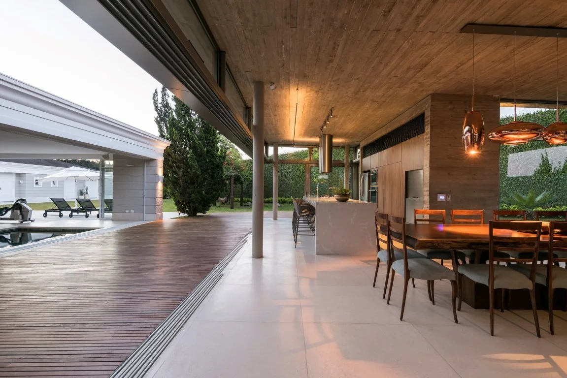 A modern indoor-outdoor dining space features a live-edge wooden table, a kitchen island, and a concrete ceiling opening to a wooden deck.