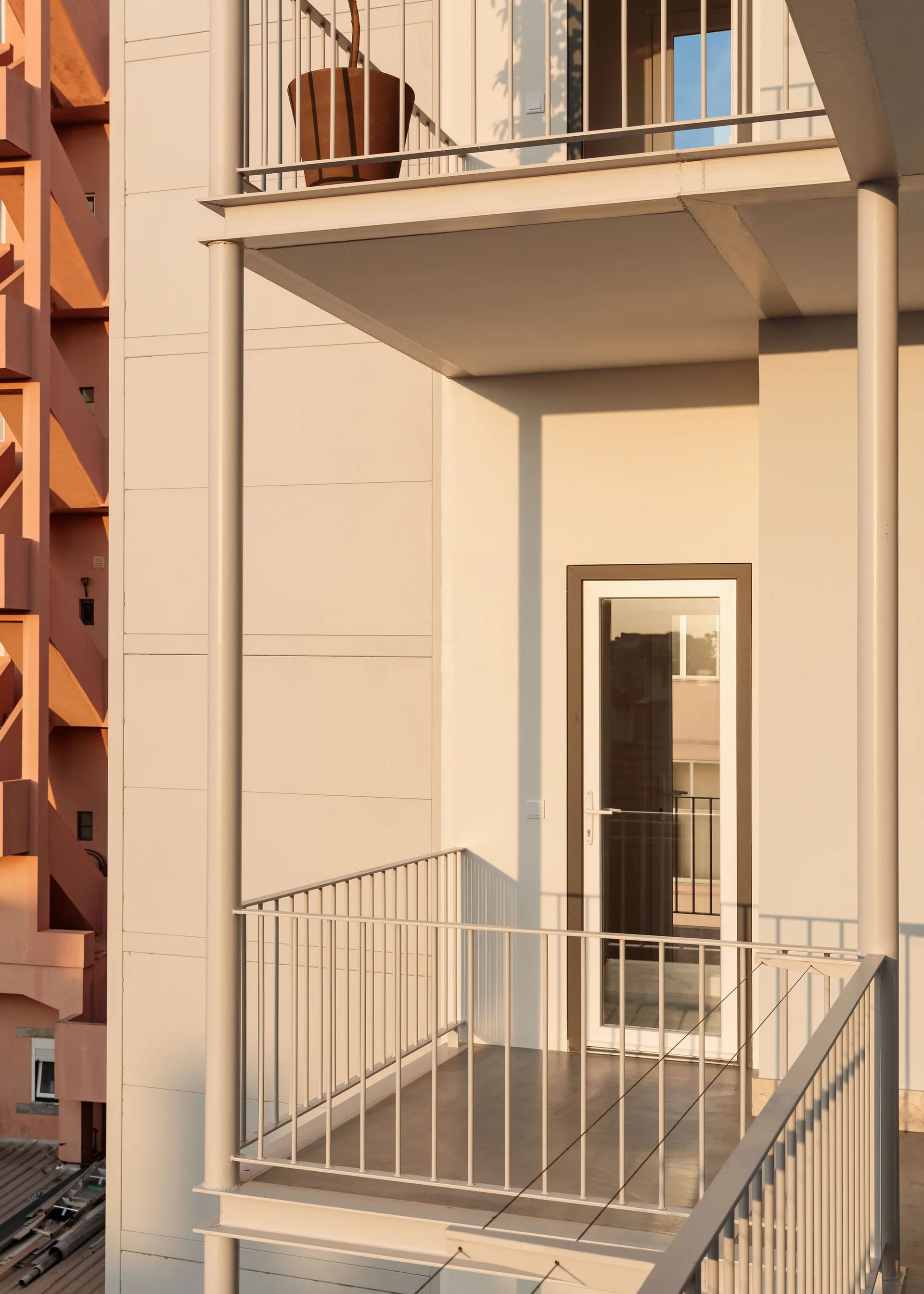 A minimalist apartment balcony featuring warm beige concrete walls, a slim metal railing, a glass door, and a brown planter in bright sunlight.