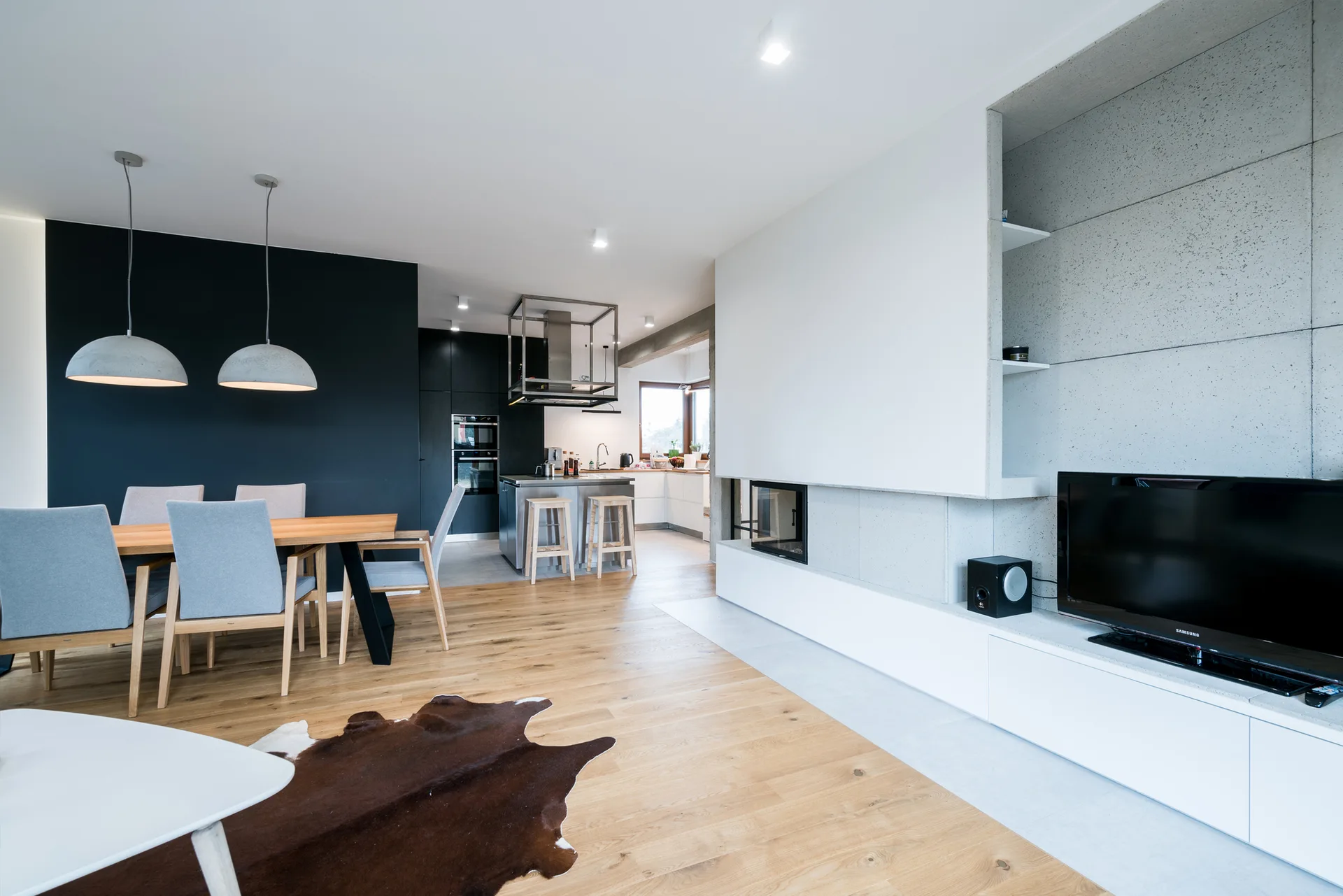 A modern open-plan living and dining room with a concrete-look media wall, a wooden dining table under dome pendant lights, and an animal hide rug.