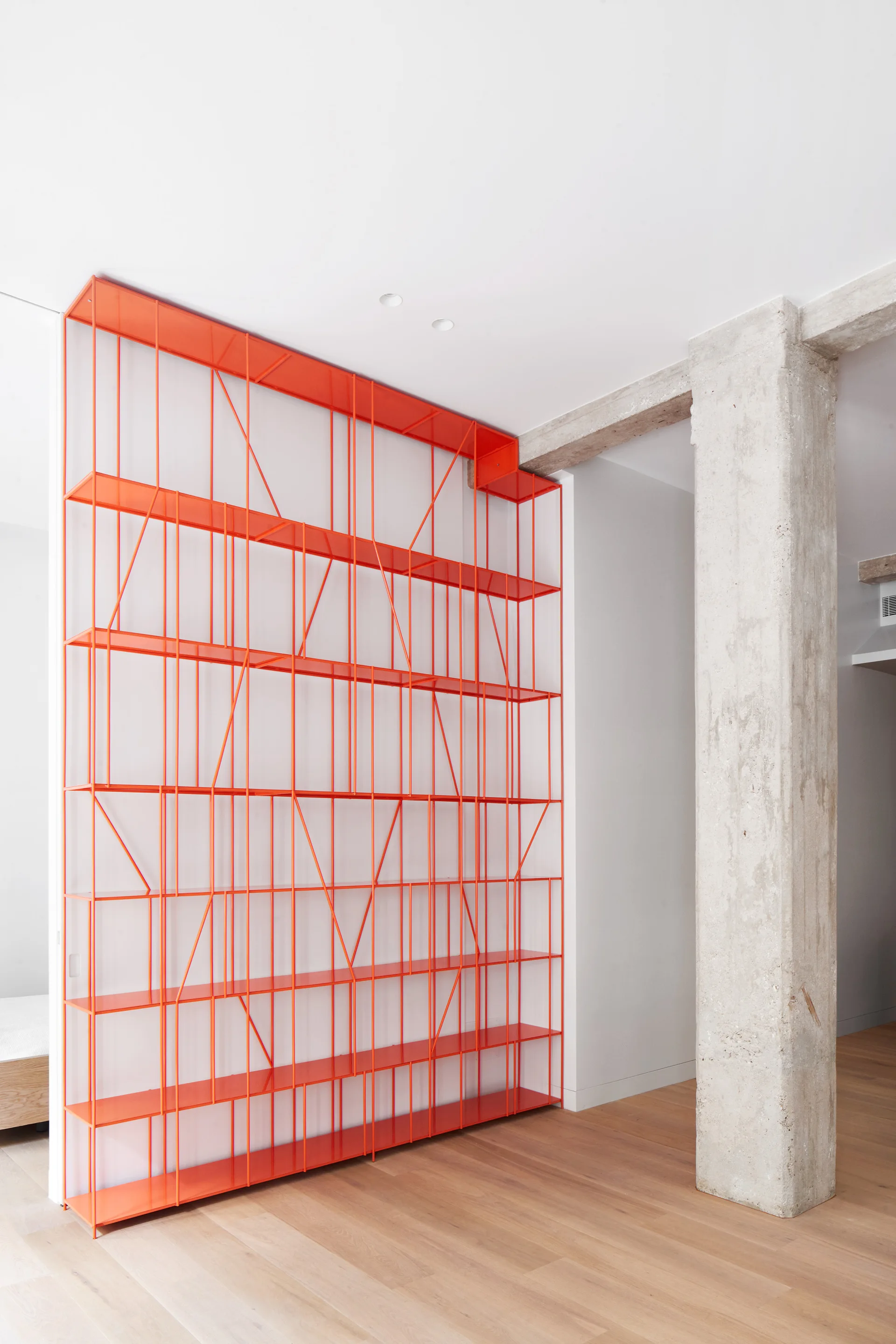 A tall modular shelving unit in bright orange metal stands against a white wall next to a rough concrete column.