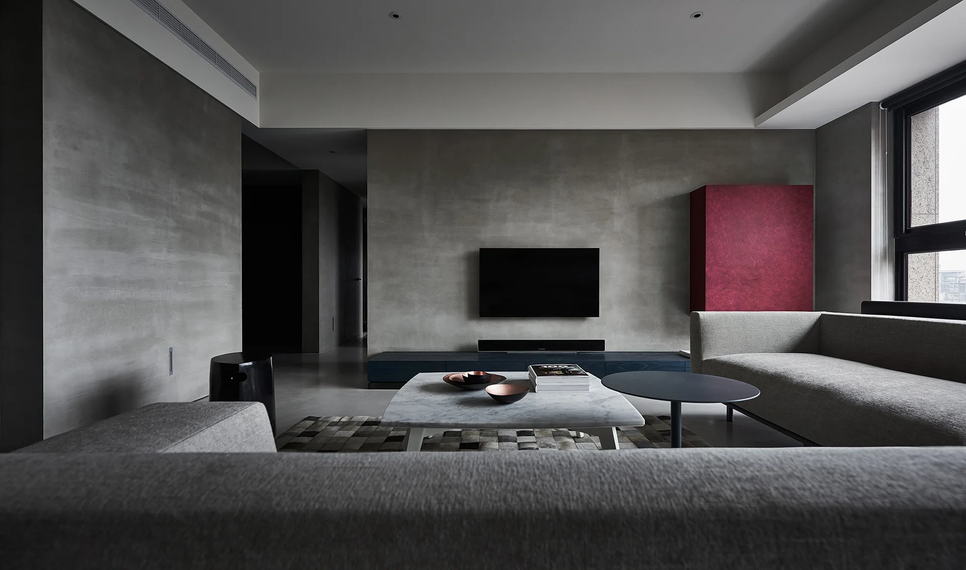 A modern minimalist living room featuring textured gray concrete walls, gray sofas, a white marble coffee table, and a bright red floating cabinet.