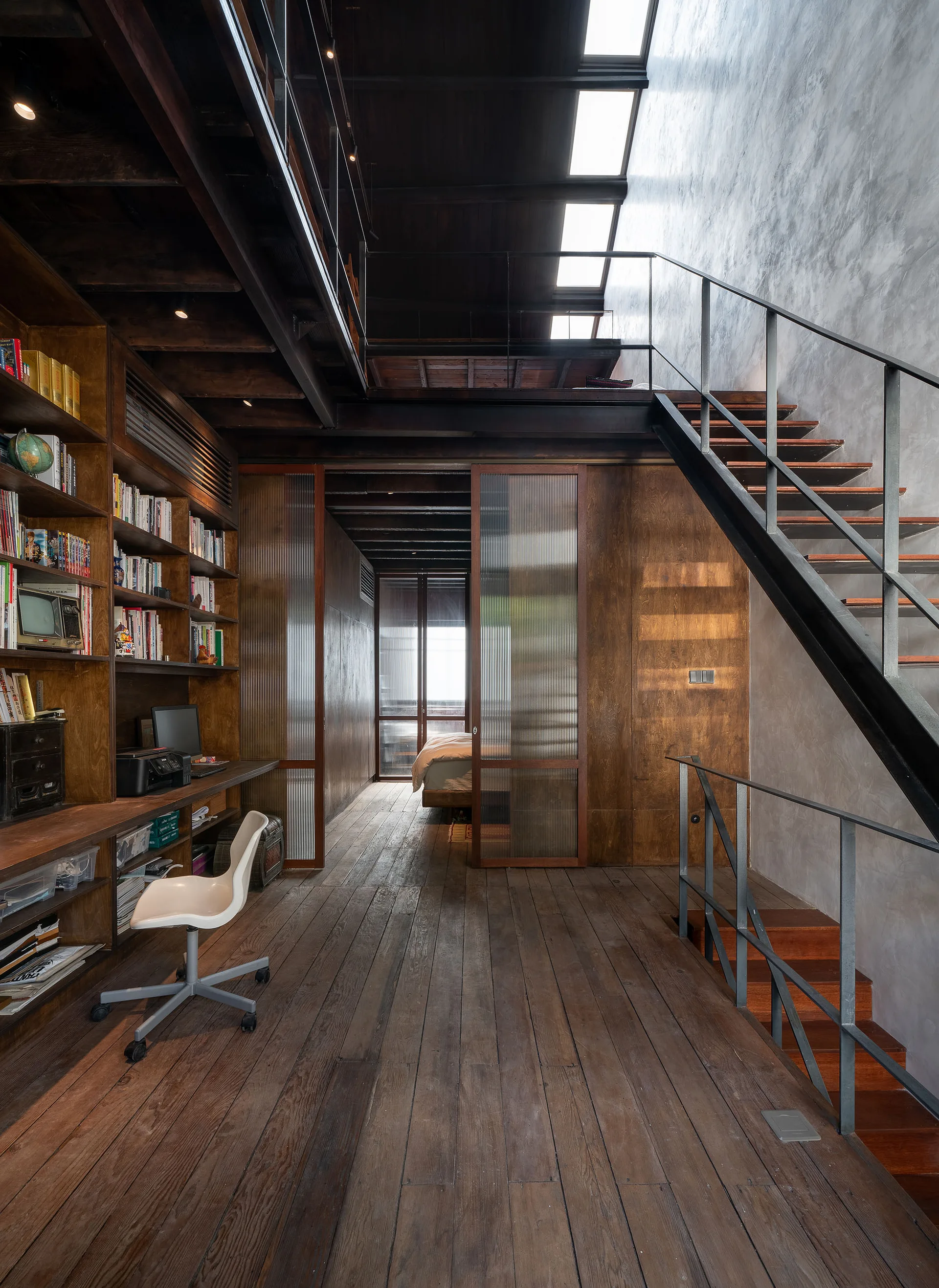 A rustic home office features a tall wooden bookshelf, a metal staircase, and sliding fluted glass doors leading to a bedroom.