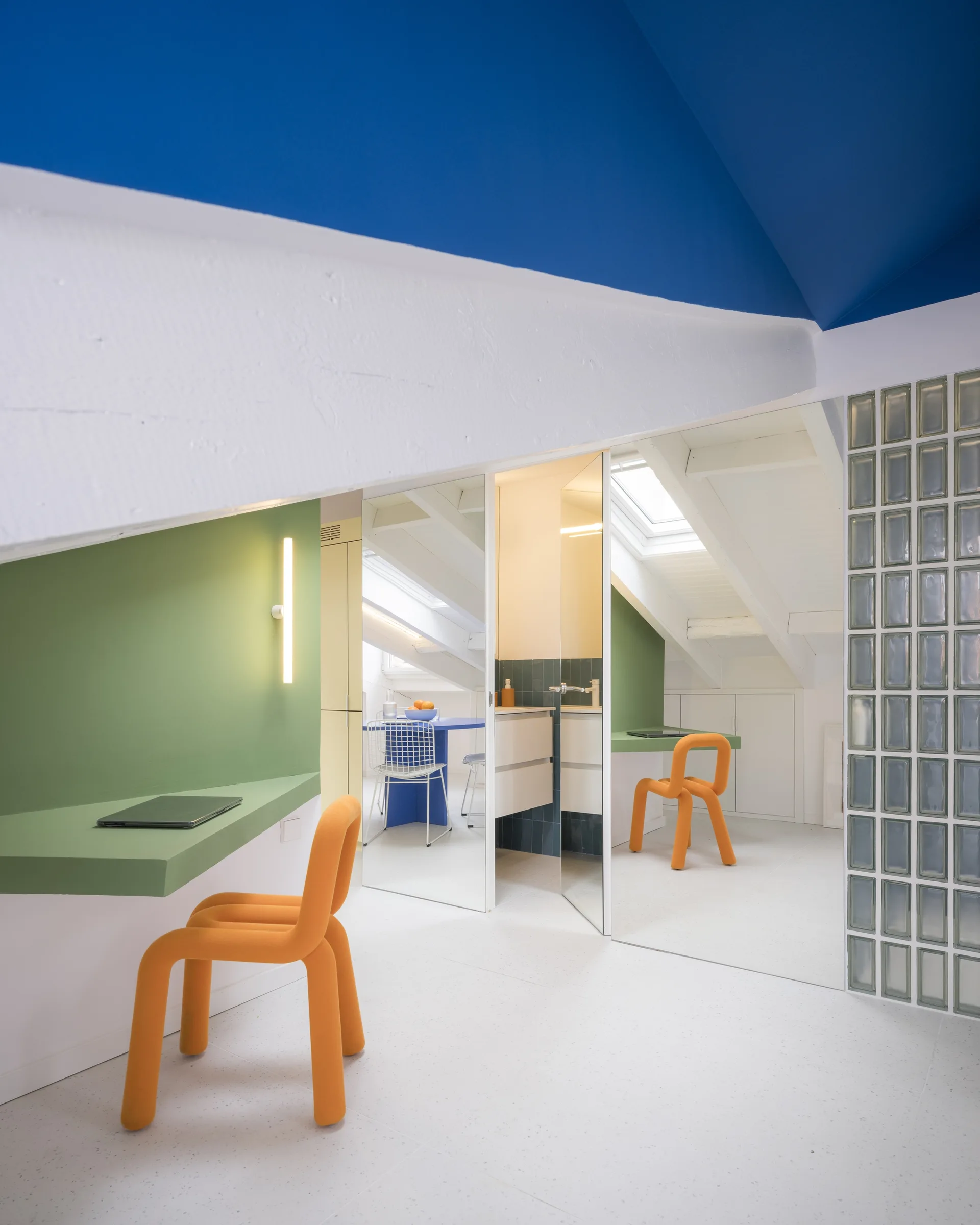 A modern attic workspace features a green floating desk, a sculptural orange chair, and a mirrored partition wall reflecting an adjacent room.