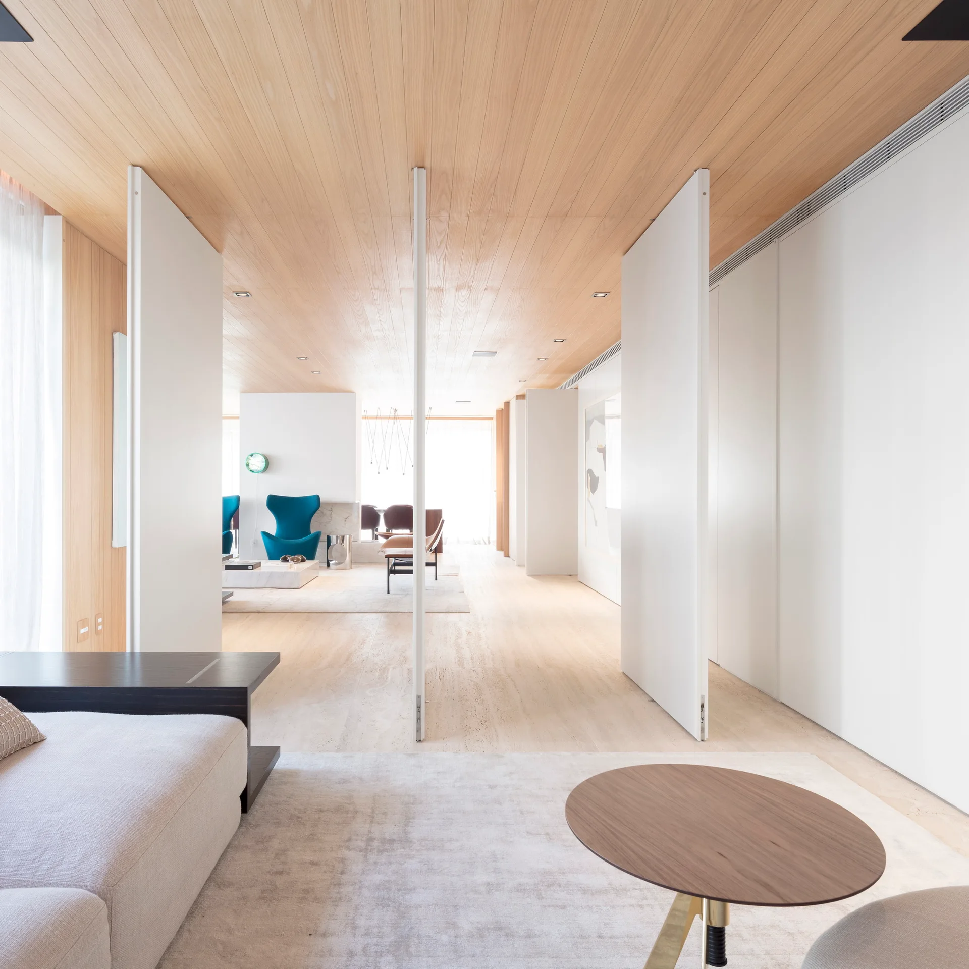A minimalist living space featuring a light wood plank ceiling, tall white pivoting partition walls, and a neutral sofa on a beige rug.