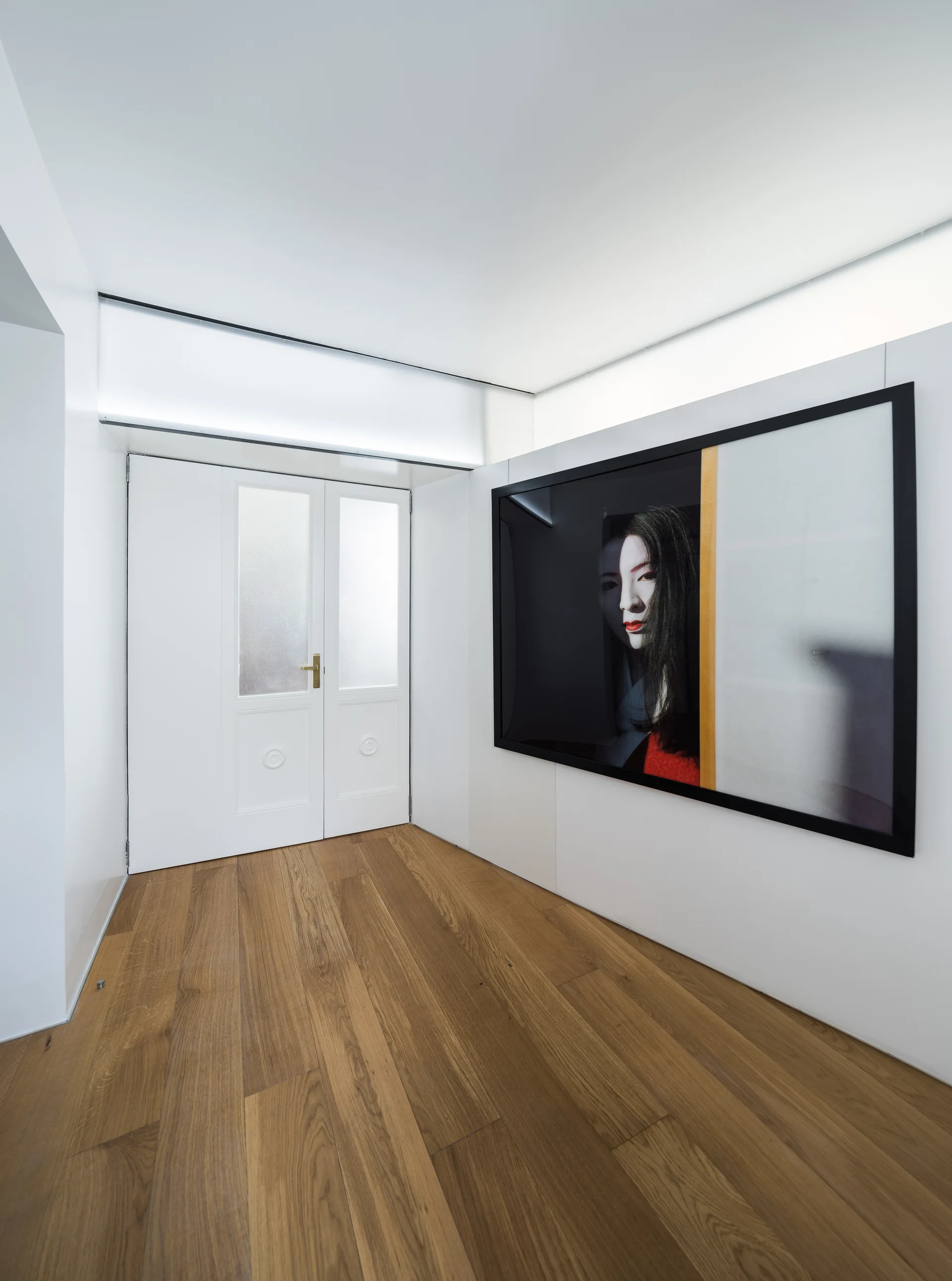 A minimalist white hallway features natural hardwood floors, a double door with frosted glass, and a large framed portrait on the wall.