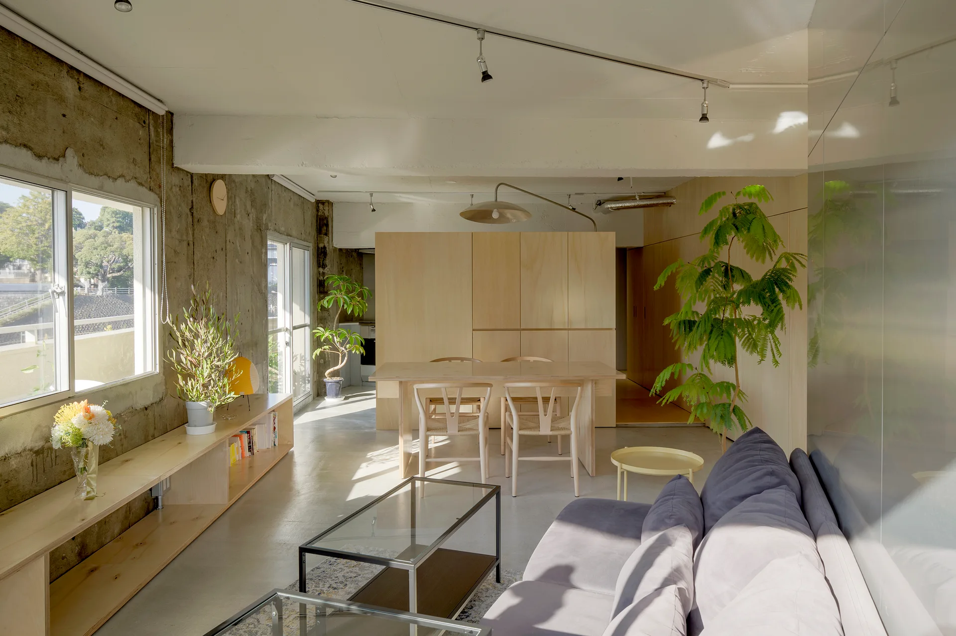A bright open-plan living and dining room with raw concrete walls, a light wood dining table with wishbone chairs, and a grey sofa.