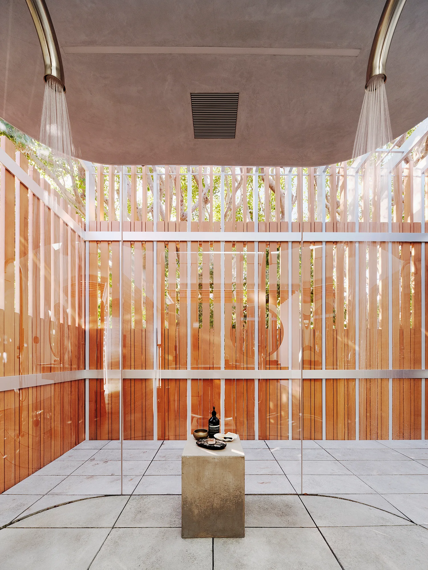 A minimalist outdoor shower featuring vertical wooden slat screens, a gray concrete tiled floor, and two curved metal shower heads pouring water.
