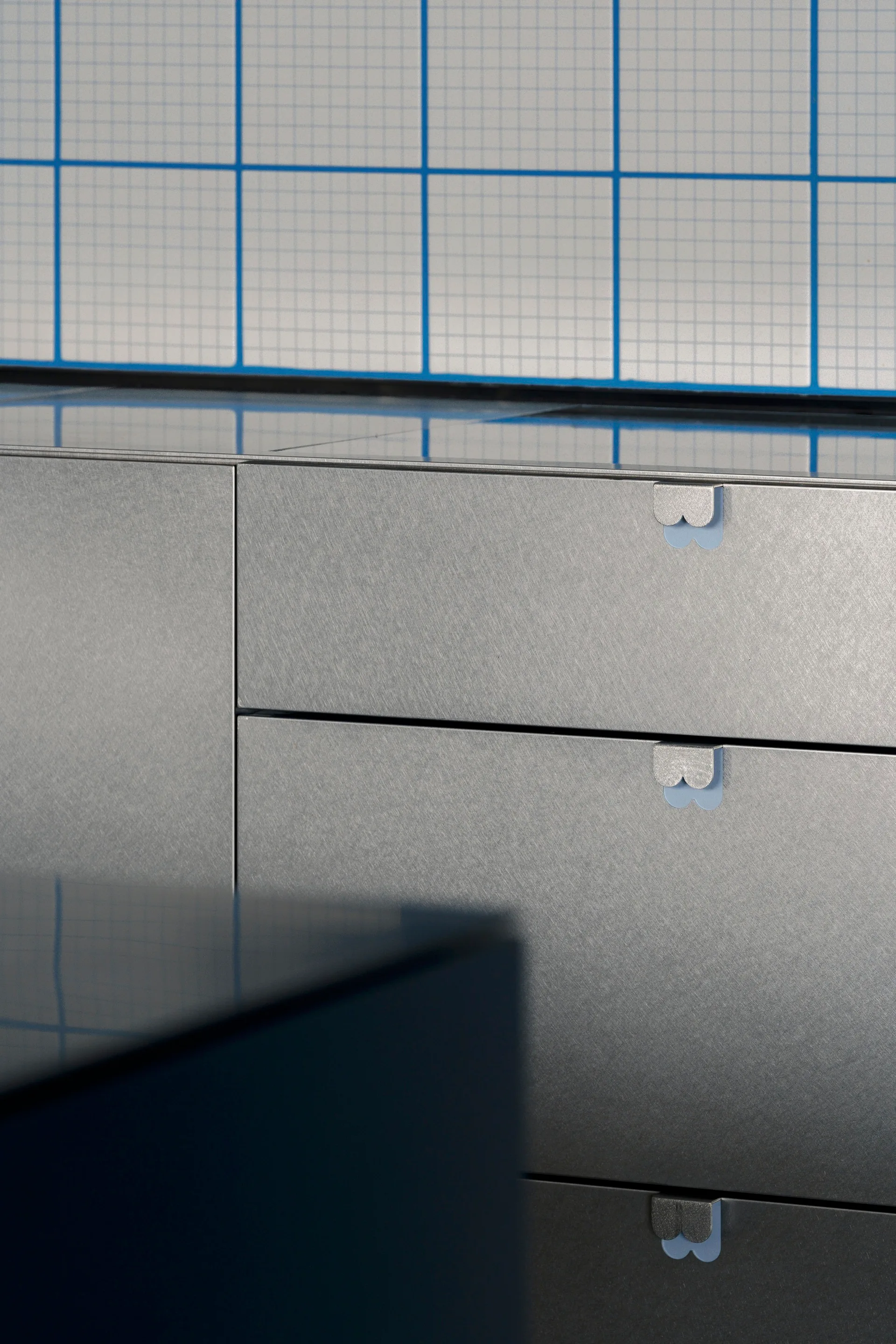 Close-up of brushed metal drawers with scalloped silver pulls set against a white wall tile with a thin blue grid pattern.