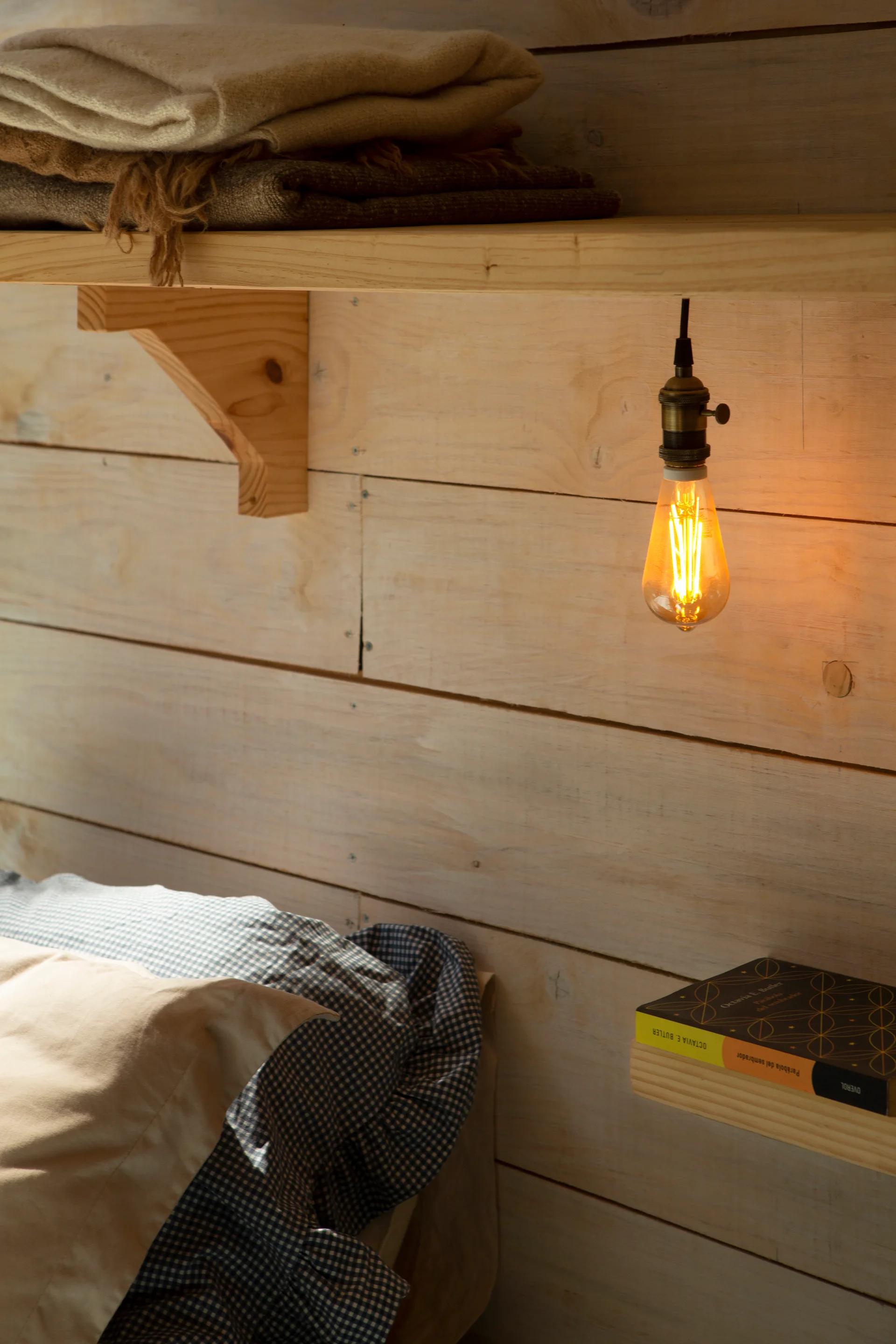 A rustic bedroom wall features horizontal wood planks, a floating shelf with folded blankets, a hanging filament bulb, and gingham pillows.