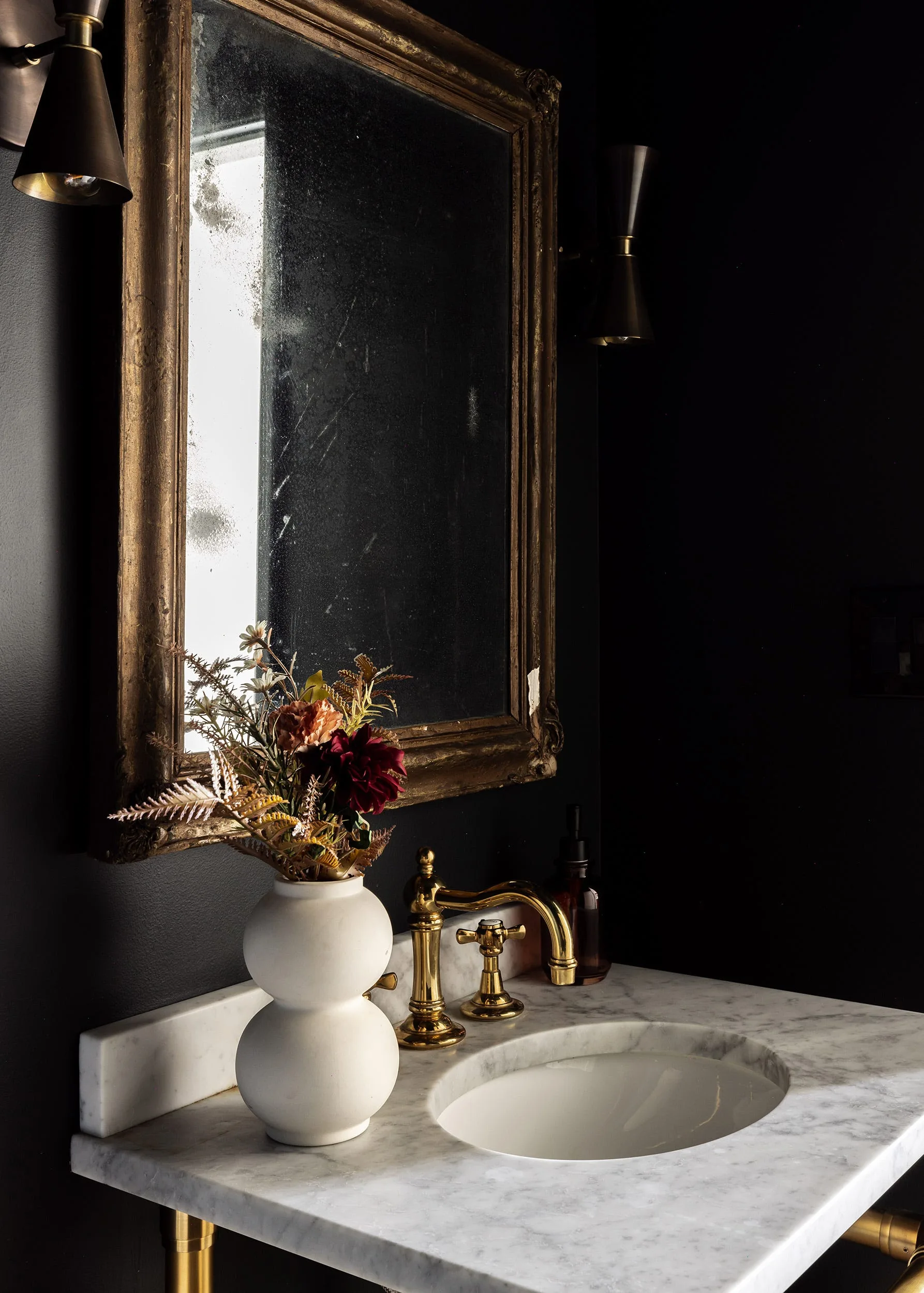 A bathroom vanity featuring a white marble countertop, a polished gold bridge faucet, and an antique gold-framed mirror against matte black walls.
